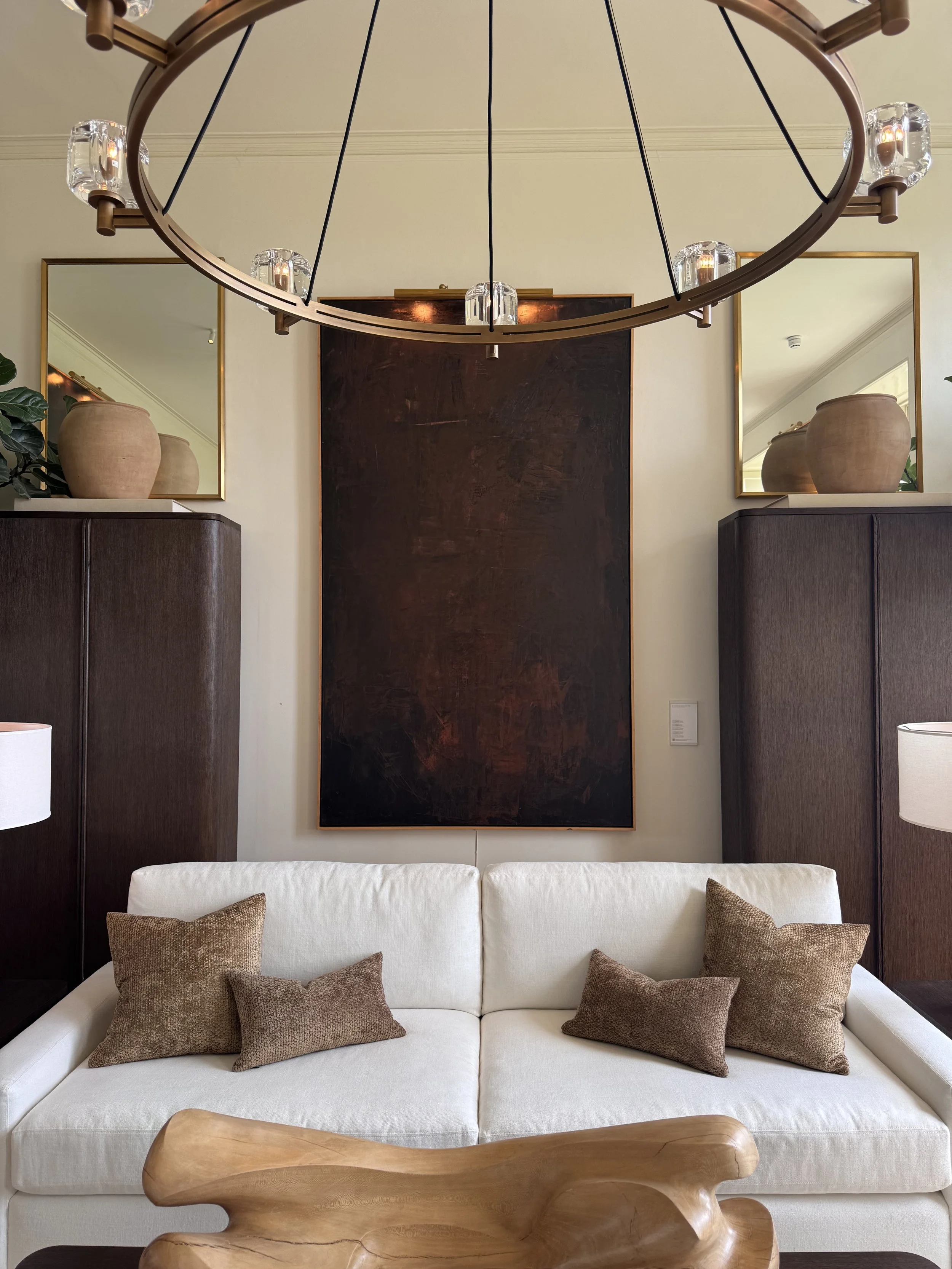 A white sofa with brown throw pillows in a living room. Behind the sofa are two large dark wood cabinets, with a large dark artwork between them. On top of each cabinet is a large beige clay pot. Two large mirrors are mounted on the wall, reflecting part of the room. A modern circular chandelier hangs above, with multiple glass candle holders.