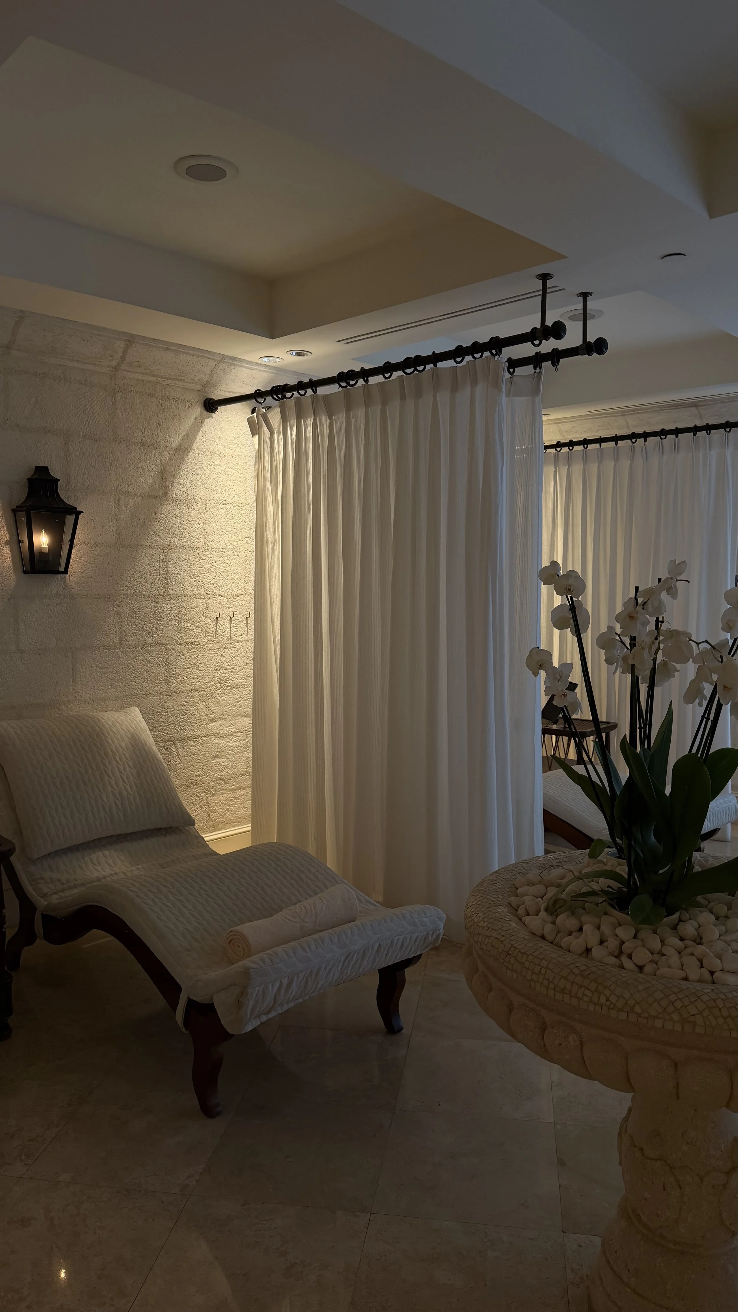 A cozy indoor space with beige tiled flooring, a lounge chair with a rolled towel, a stone wall with a black lantern-style wall light, and a sheer curtain dividing the space. There is a large orchid plant with white flowers in a stone planter and a small wooden table in the background.