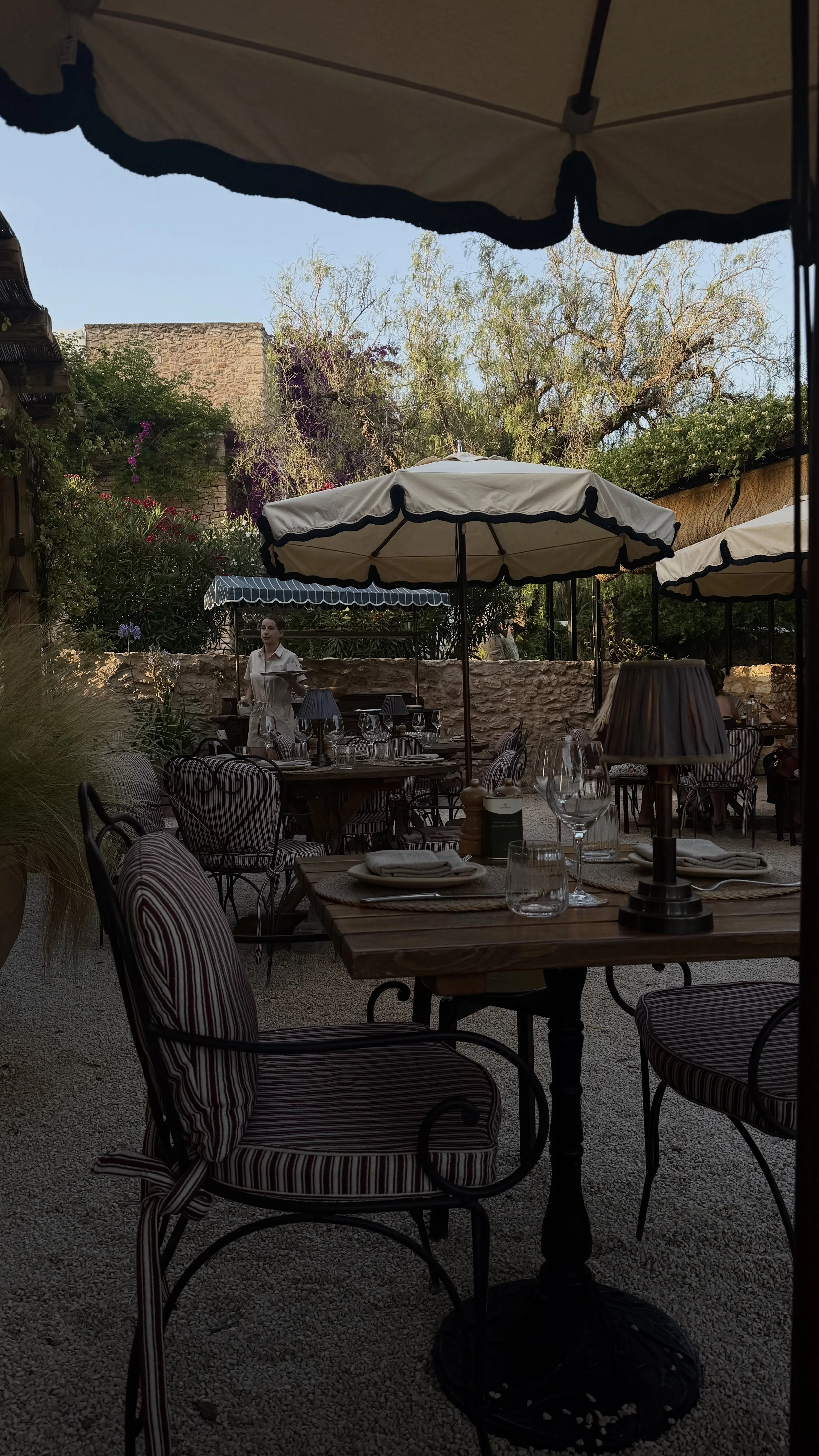 Outdoor restaurant patio with striped cushioned chairs, tables set with glasses, plates, and cutlery, under large white umbrellas, surrounded by trees and greenery.