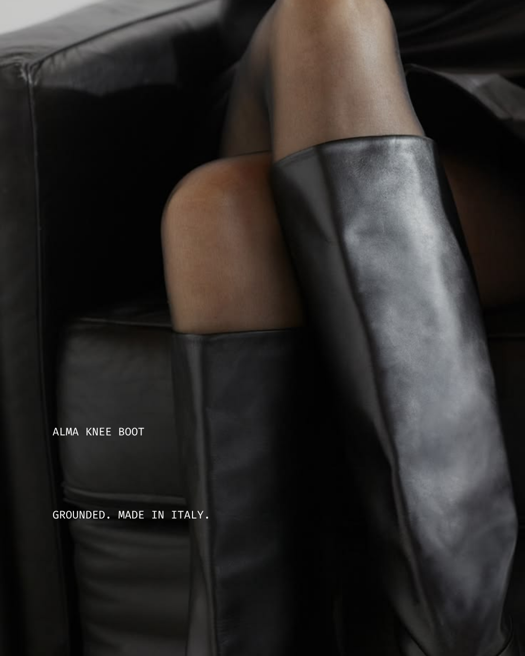 A person sitting on a black leather sofa wearing a black knee-high leather boot and sheer black stockings. The photo is close-up, with text overlay reading 'ALMA KNEE BOOT GROUND. MADE IN ITALY.'
