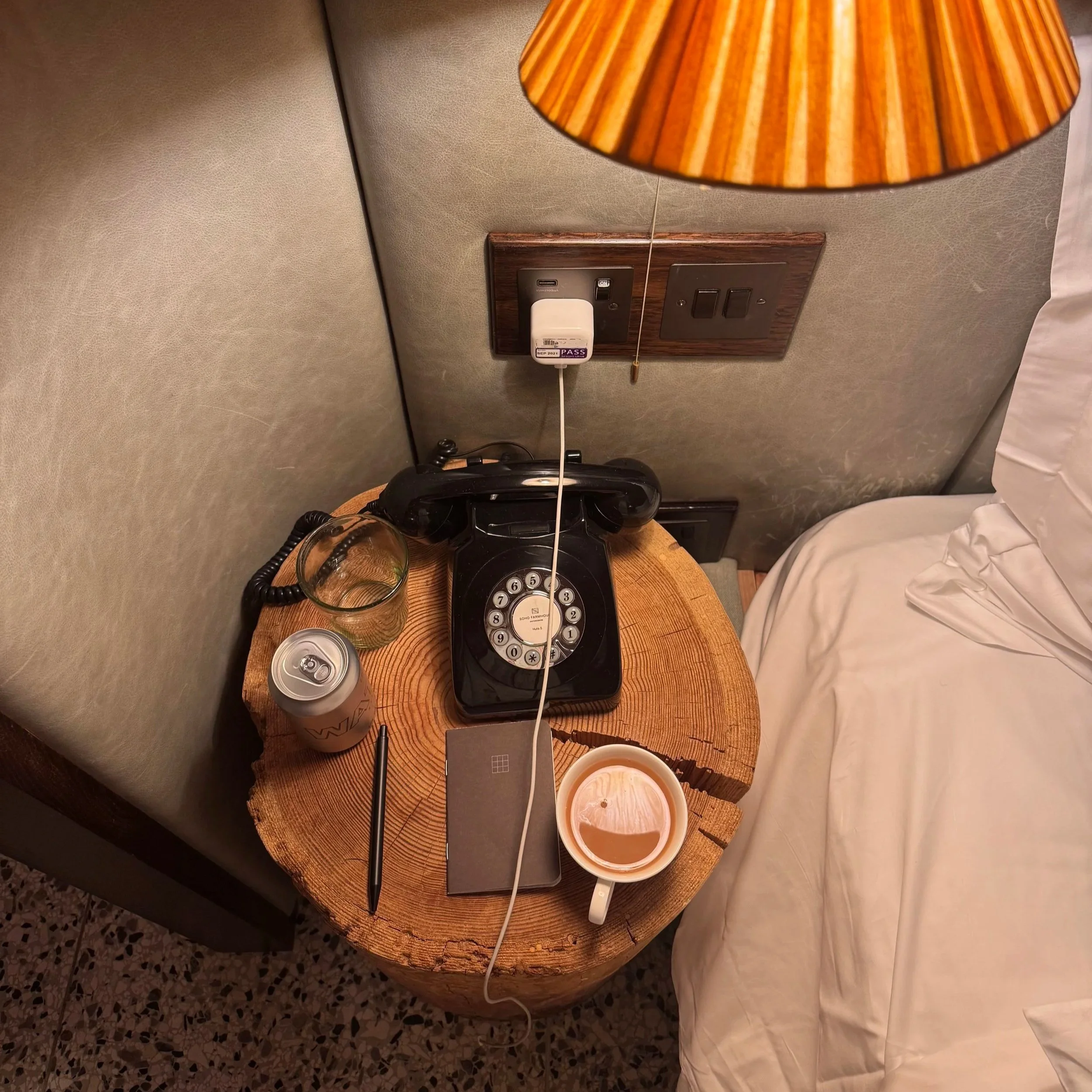 Close-up of a nightstand with a lamp, an old-fashioned rotary phone, a glass of water, a USB wall charger, a can, and some cups on a wooden surface. Part of a bed with white bedding is visible to the right.