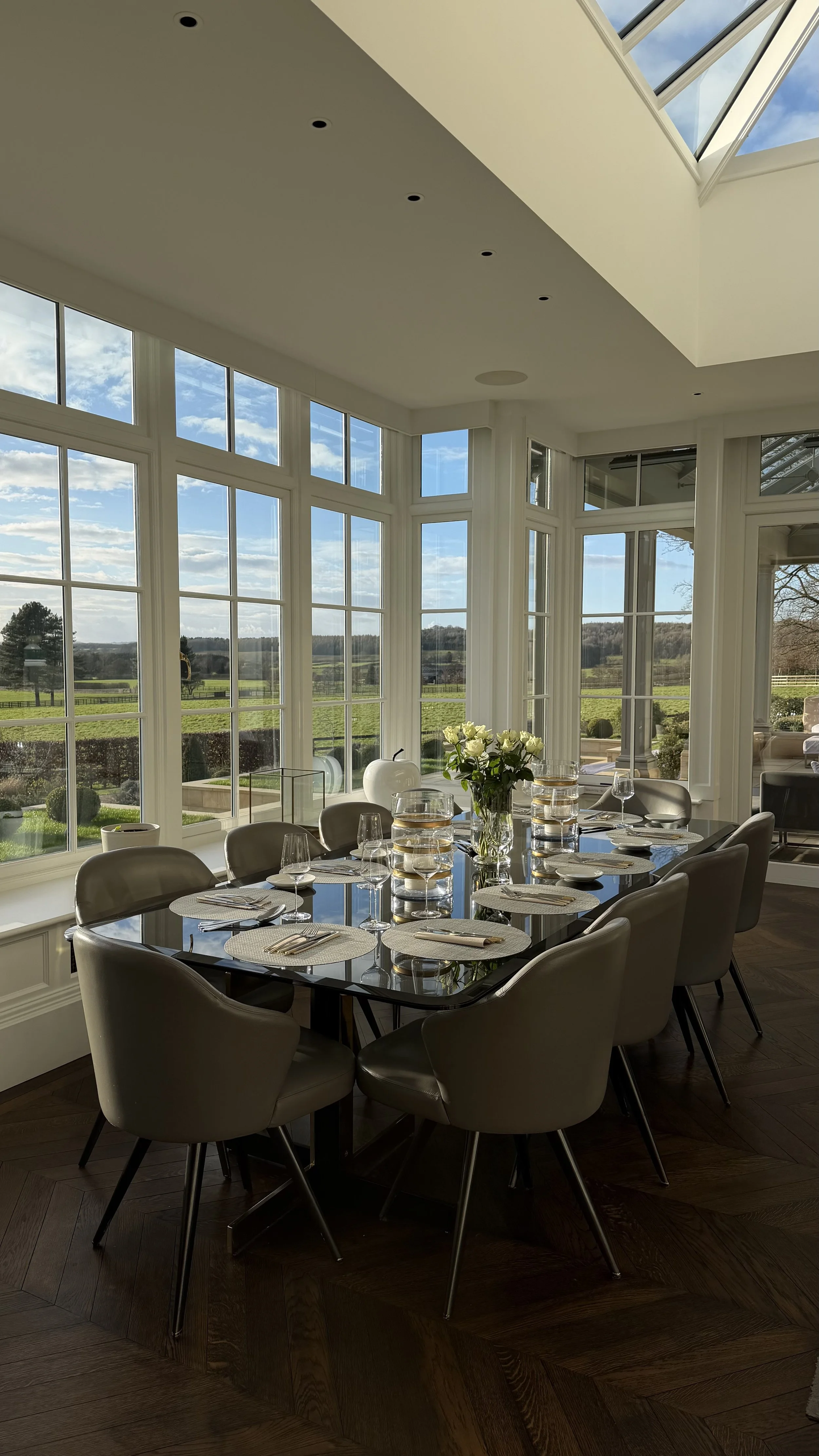 A dining room with a large glass table, set with plates, glasses, and napkins, surrounded by beige chairs. The room has large windows and a glass ceiling, offering a view of the outdoors with green fields and a blue sky.