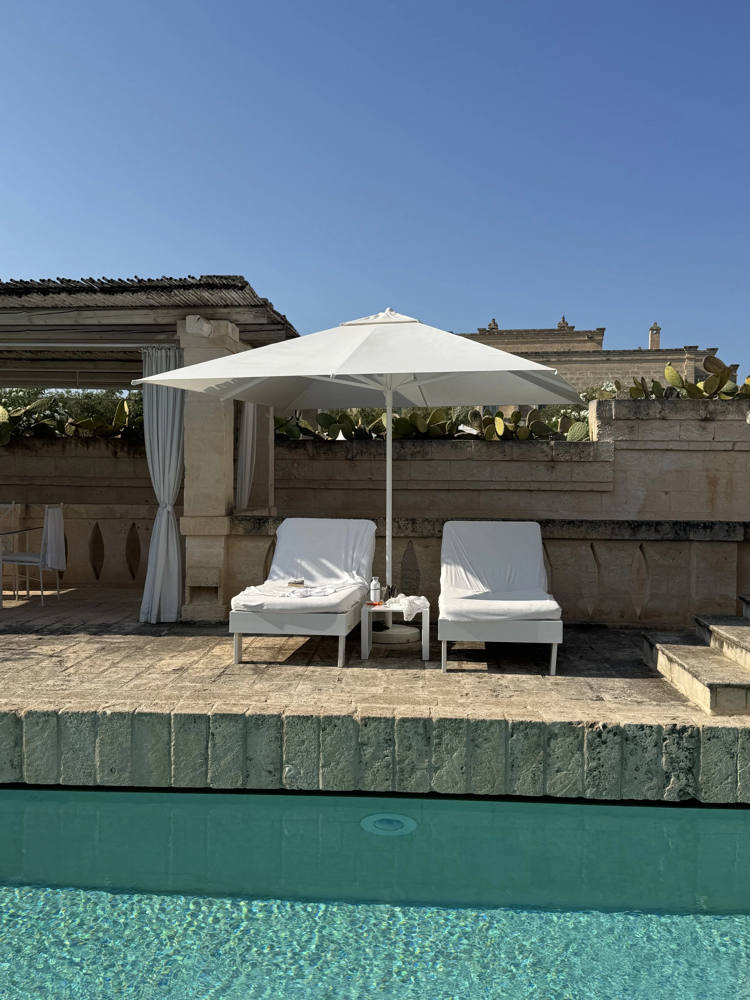 A swimming pool with blue water, white lounge chairs, and white umbrellas on a patio. A beige stone wall with cactus plants behind it and a clear blue sky overhead.