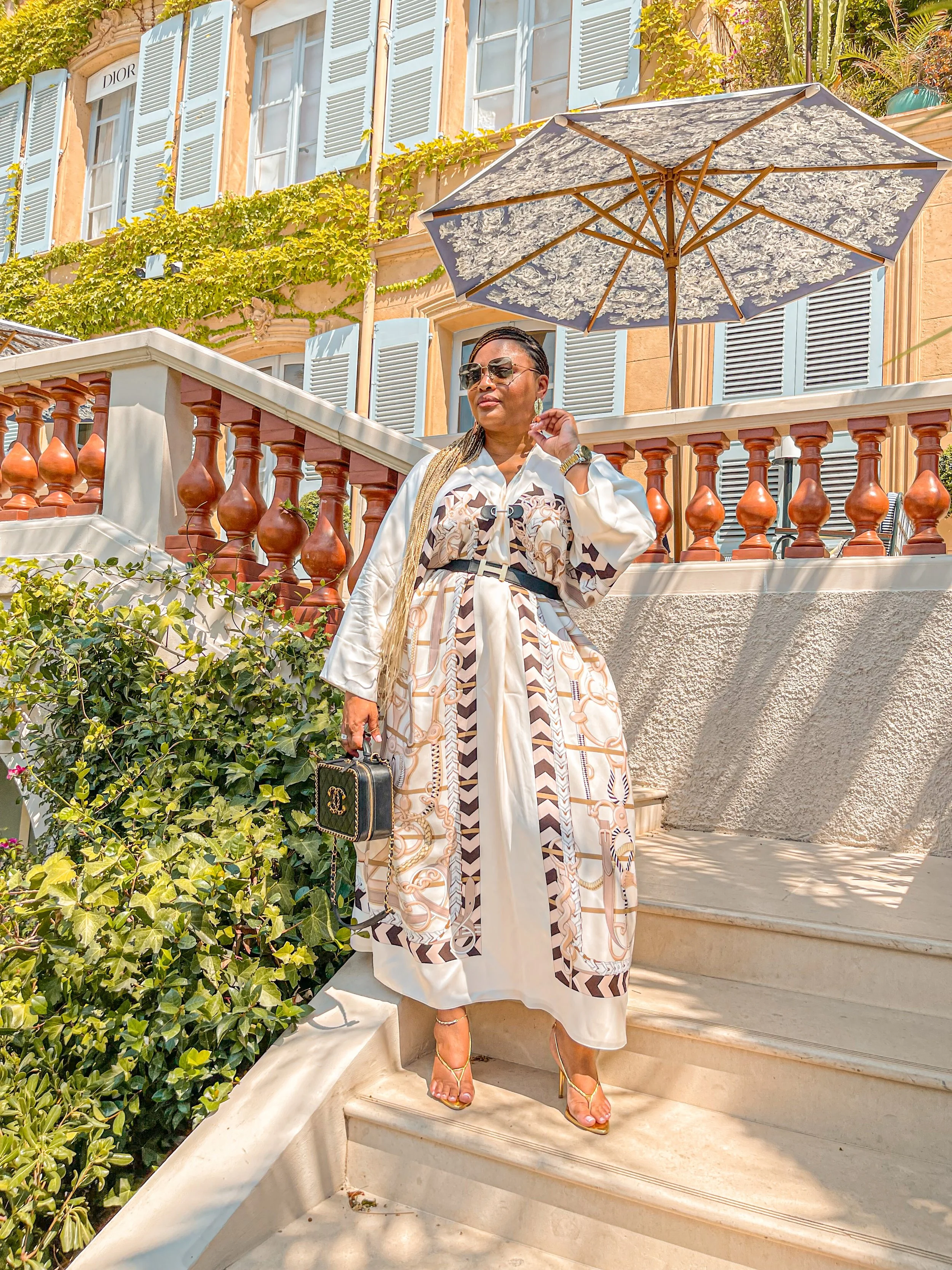 Woman in elegant Hermès dress and sunglasses poses on sunny steps with umbrella and greenery.