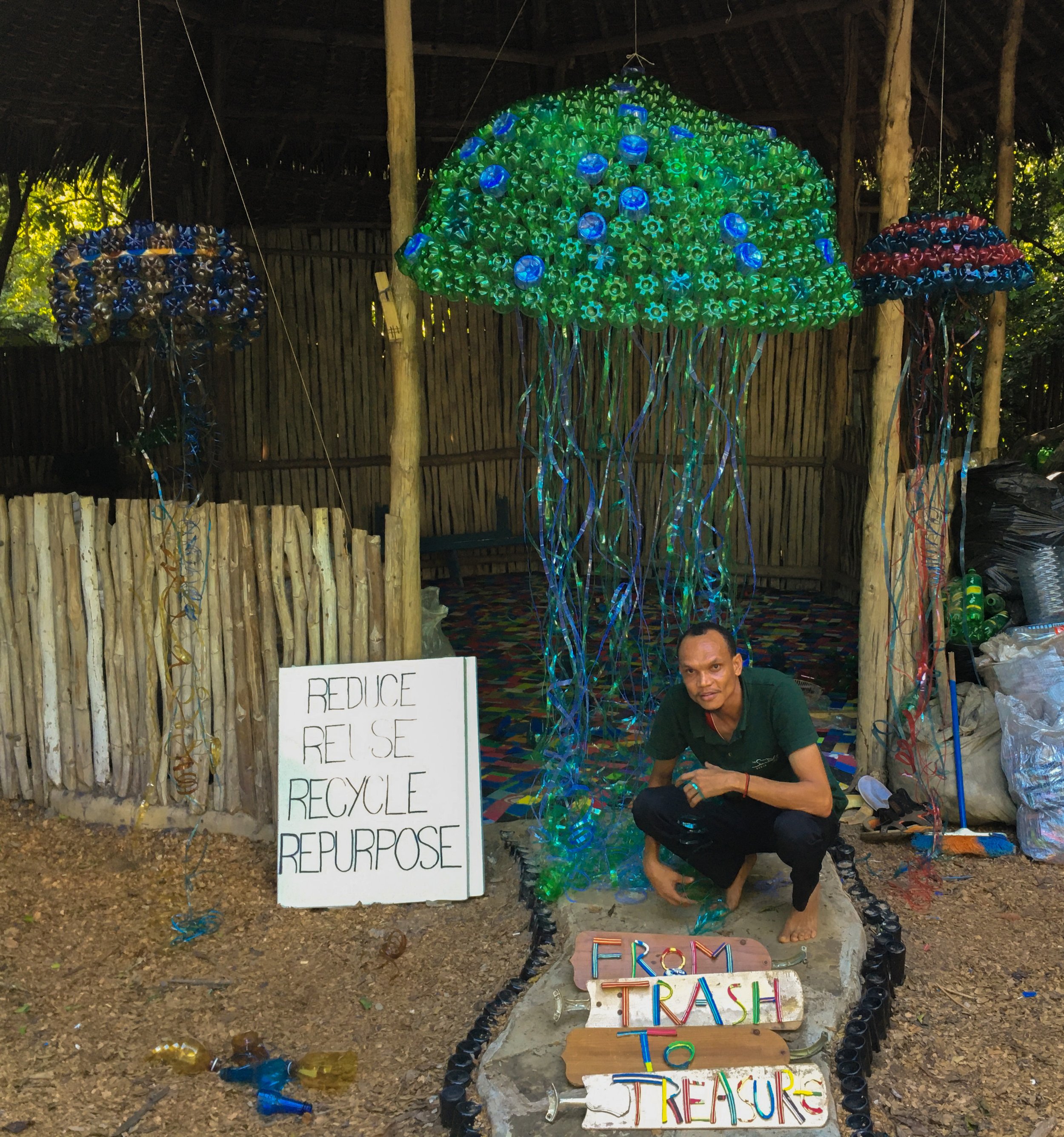 A man squatting near a large, colorful, illuminated sculpture of a crab made from plastic bottle caps. The sculpture is decorated with green, blue, and red caps. Behind him, there are signs with words about reducing, reusing, recycling, and purpose, as well as a sign with the words 'Earth Trashion' and colorful letters. The setting appears to be an outdoor area with wooden poles, a bamboo fence, and trees.