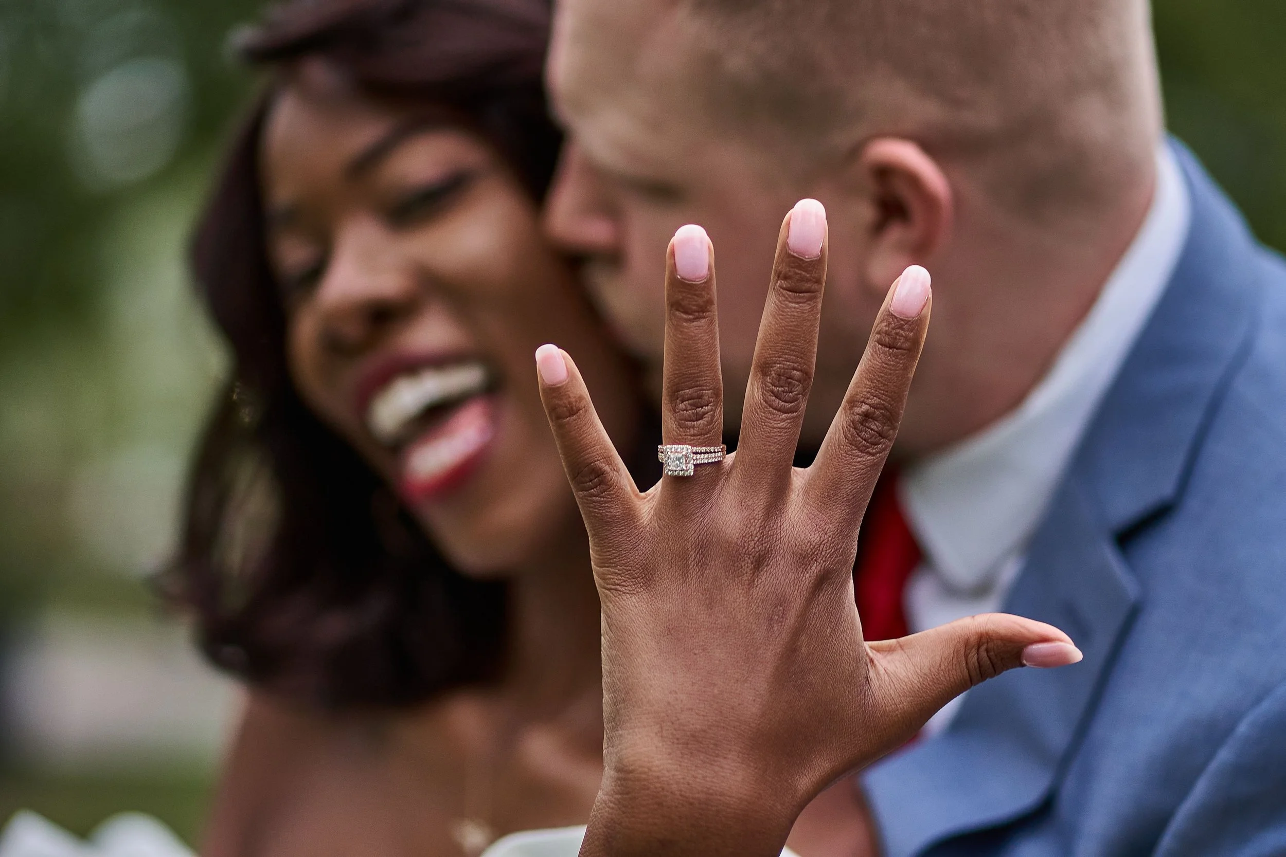 A woman smiling while holding up an engagement ring while being kissed by a man.