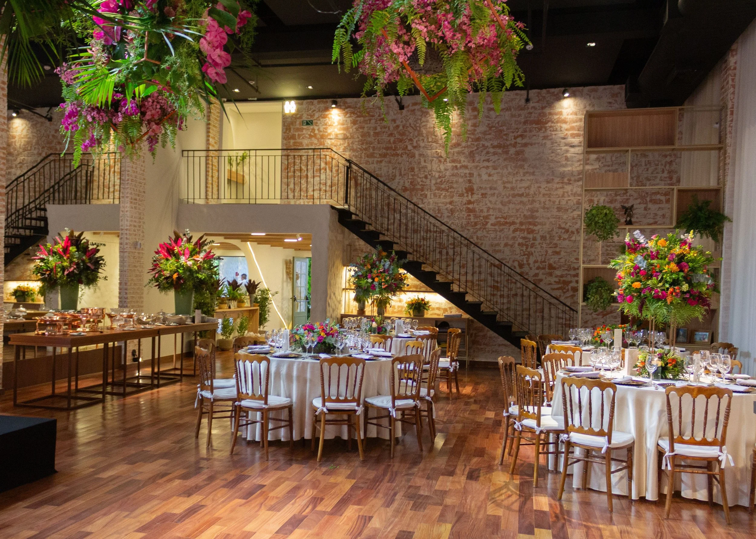 Large indoor space filled with tables and chairs decorated with white cloths, pink and green floral decorations on table and hanging from ceiling