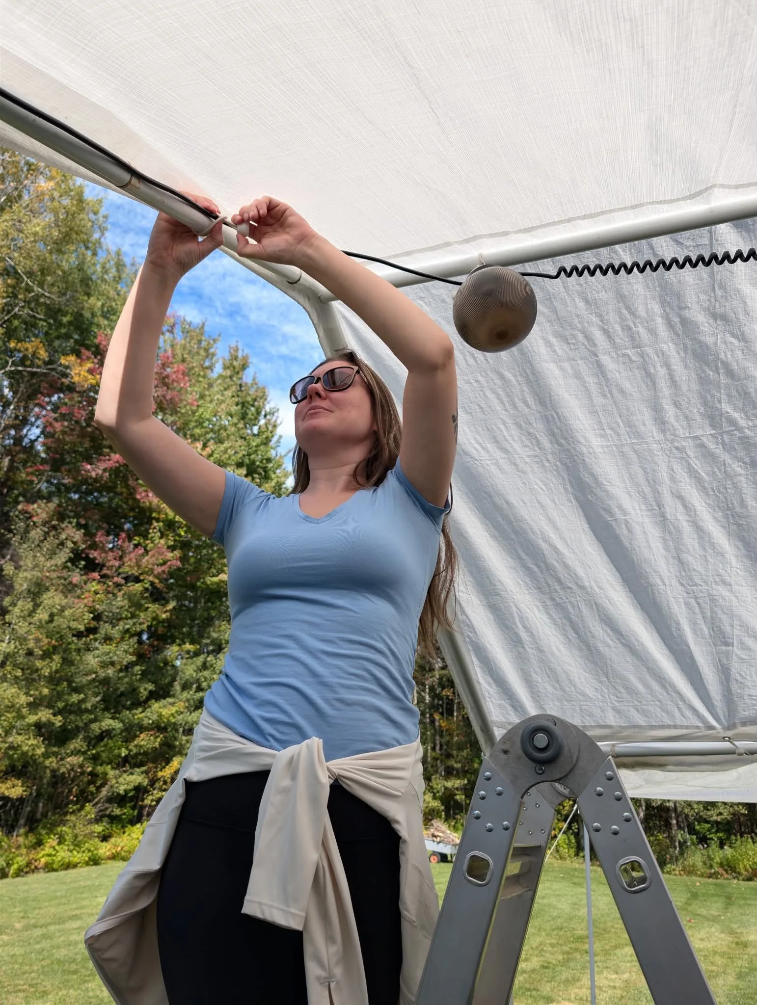 Woman in blue shirt, black pants wearing sunglasses standing on a ladder hanging large lights in a white tent with trees in the bacground.