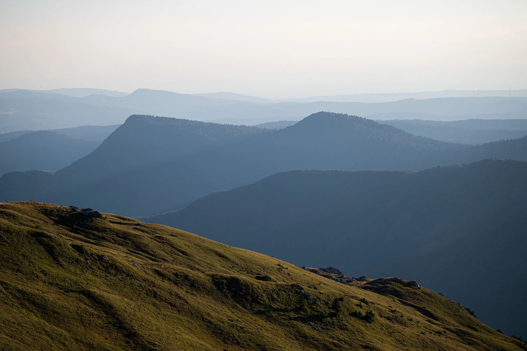 Paysage de montagnes verdoyantes avec des collines au premier plan et des montagnes plus lointaines en arrière-plan, sous un ciel clair et lumineux.