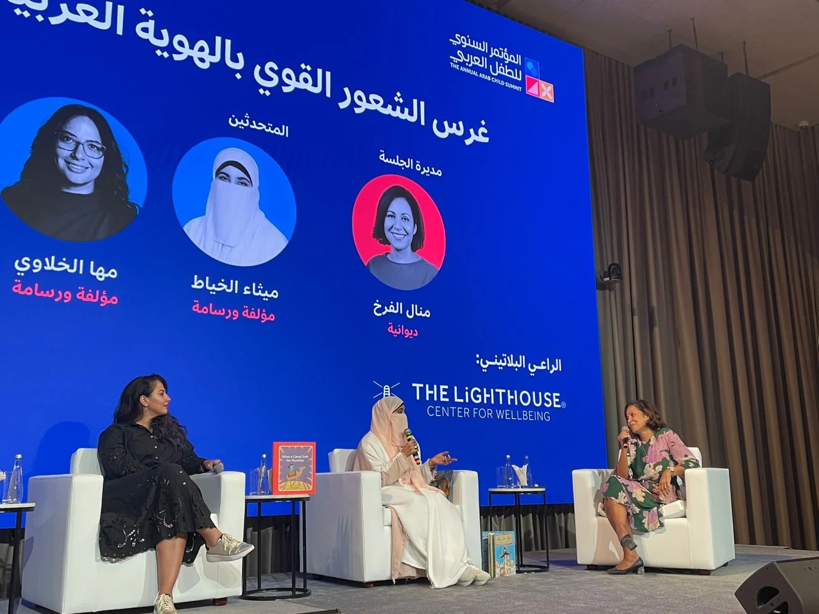 A panel discussion at the Arab Child Summit, with three women seated on stage, including one wearing a headscarf. Behind them is a large blue screen displaying the summit's logo and images of the speakers.