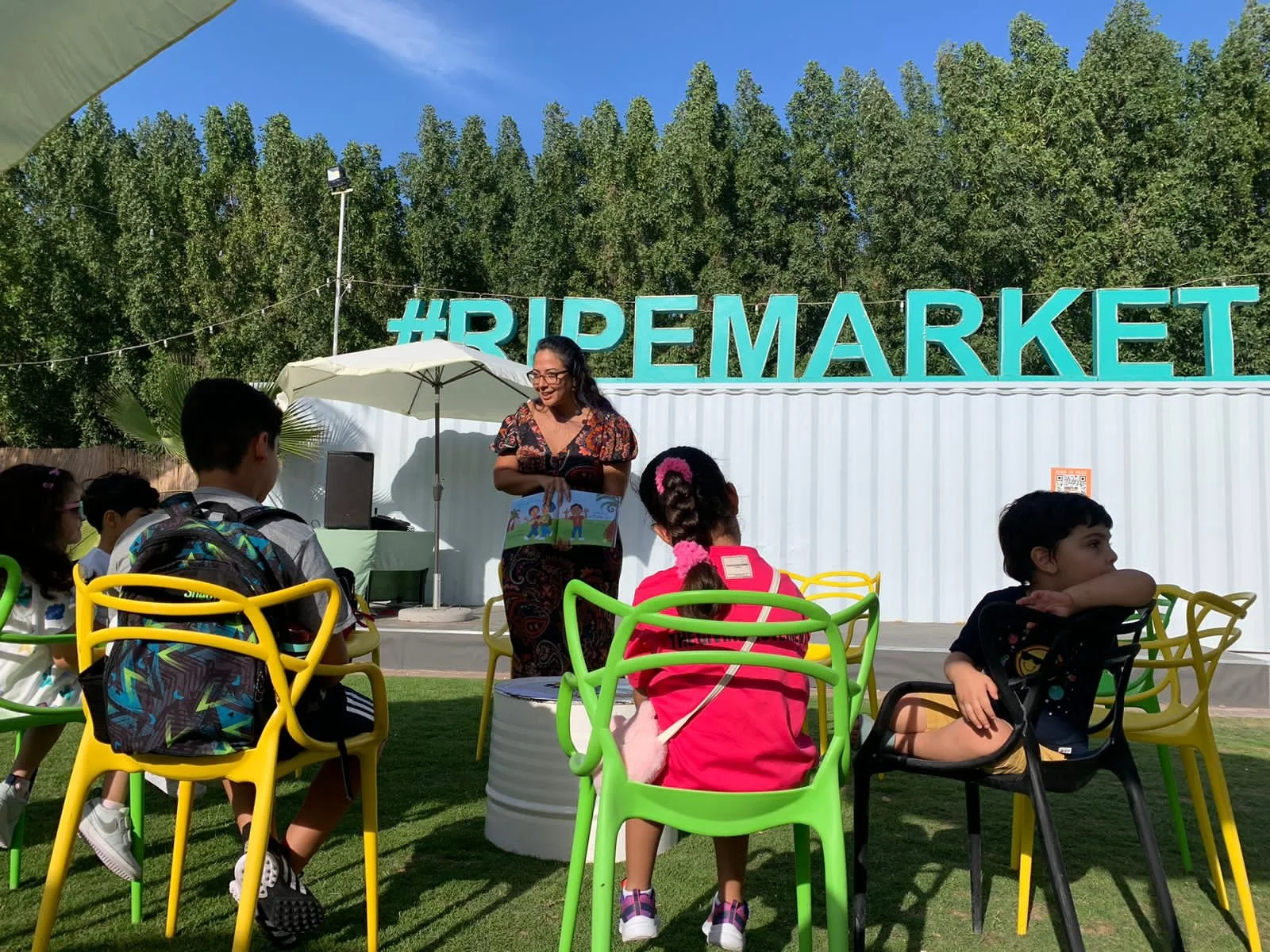 A group of children sitting on colorful chairs outdoors watching a woman read a book. There is a large hashtag sign that says '#REMARKET' in the background, with trees and a blue sky.