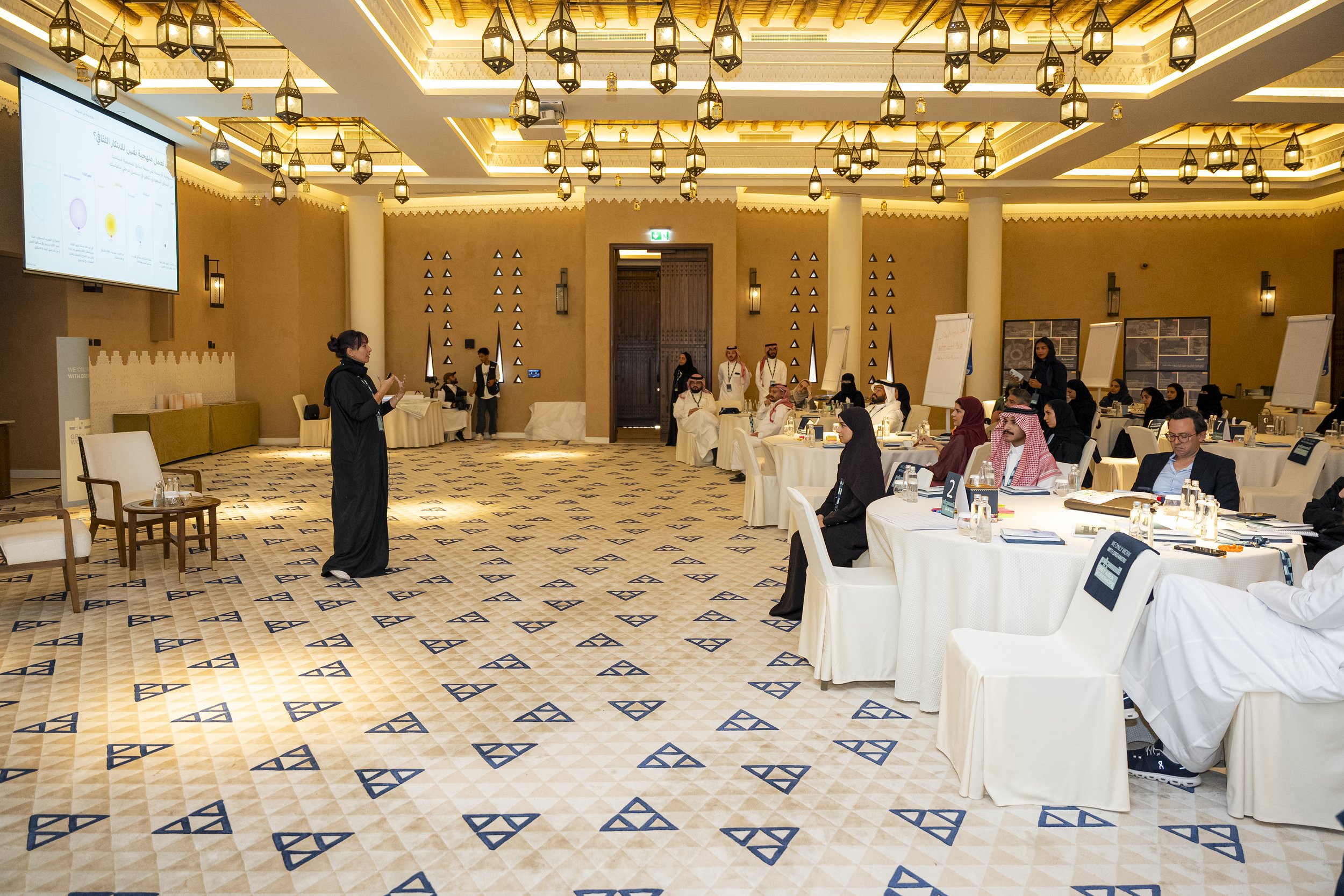 A conference room filled with people sitting at round tables covered with white tablecloths, listening to Maha in black giving a presentation and facilitating the workshop.