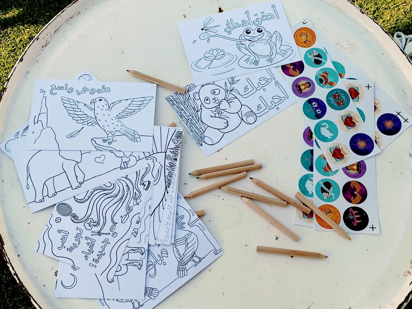 Coloring pages with animal and nature illustrations, some filled with Arabic text, displayed on a white round table with several colored pencils and sticker sheets.
