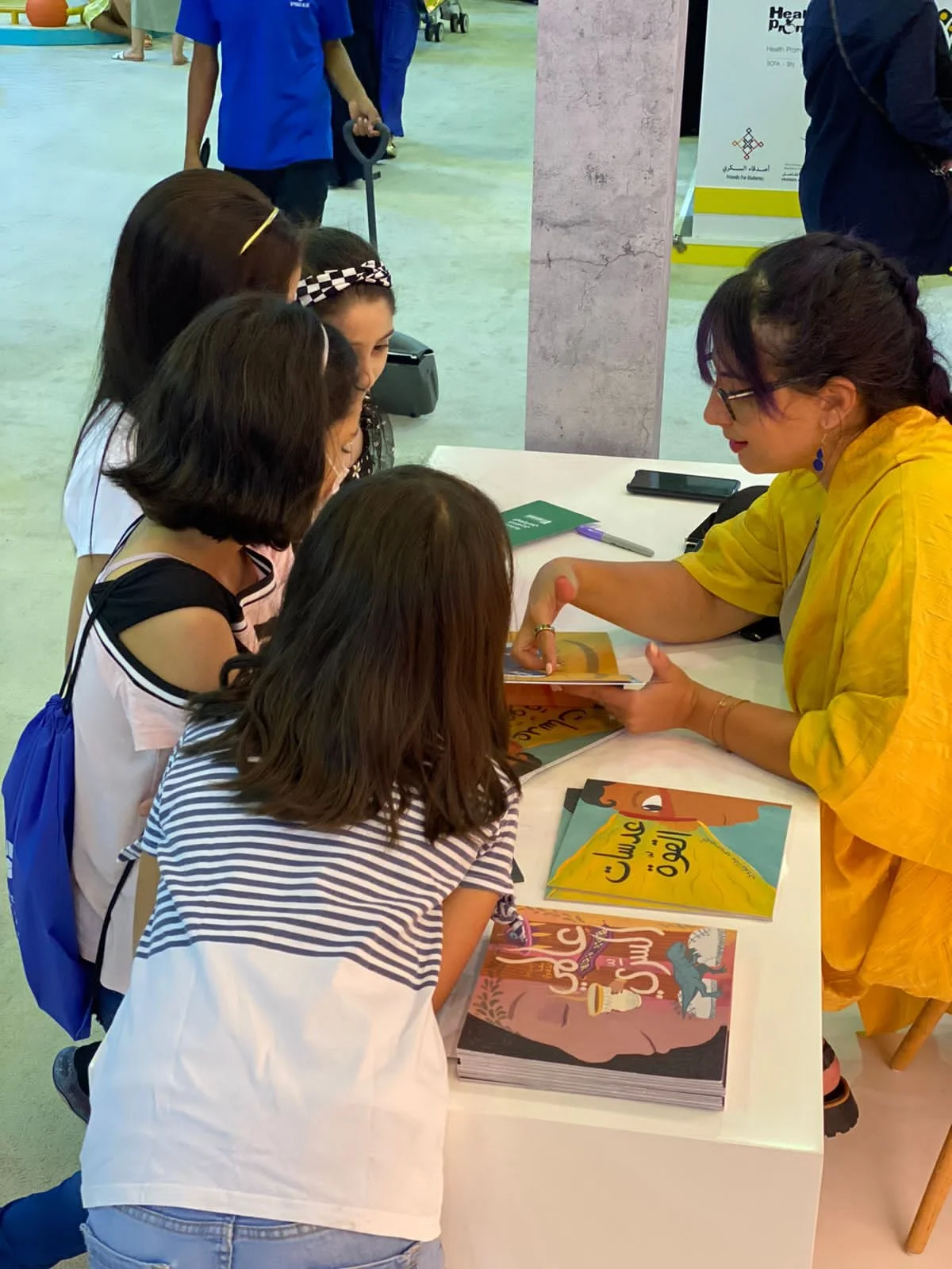 A woman in a yellow outfit sitting at a table signing books for a group of young girls at a book signing event.