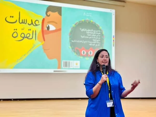 A woman speaking into a microphone in front of a projector screen displaying a colorful graphic with Arabic text and a cartoon face, in a classroom or conference setting.