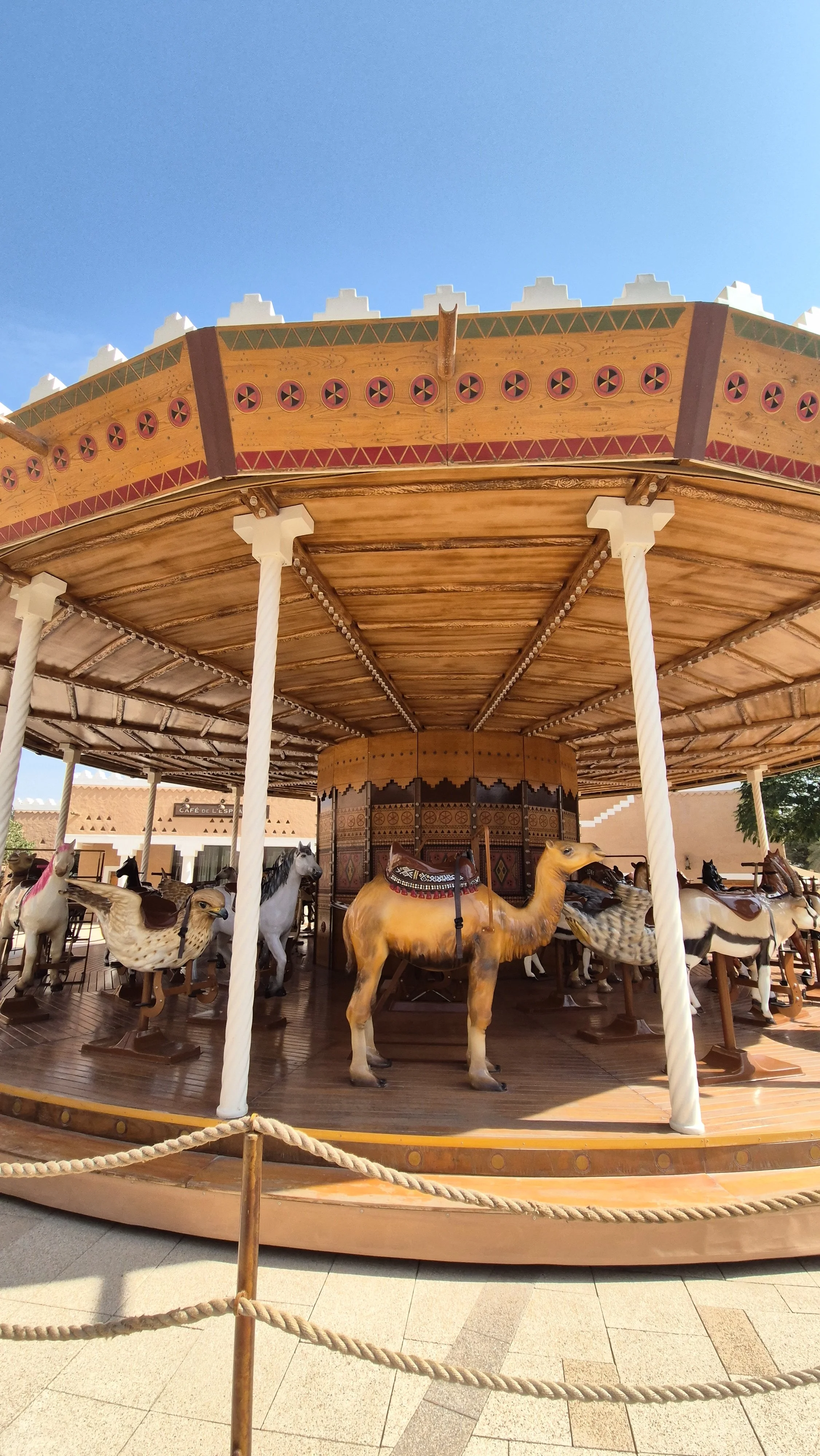 A vintage carousel with horse and camel figures, surrounded by a rope barrier, outdoors against a clear blue sky.
