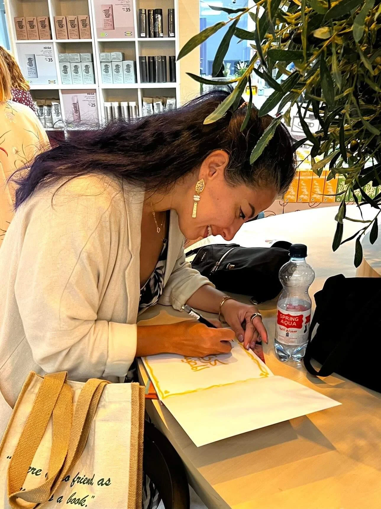 A woman with long dark hair, wearing earrings and a light-colored blazer, is seated at a table and writing on a large piece of paper. The table has a water bottle, a bag, and some papers. In the background, shelves with boxes and products are visible