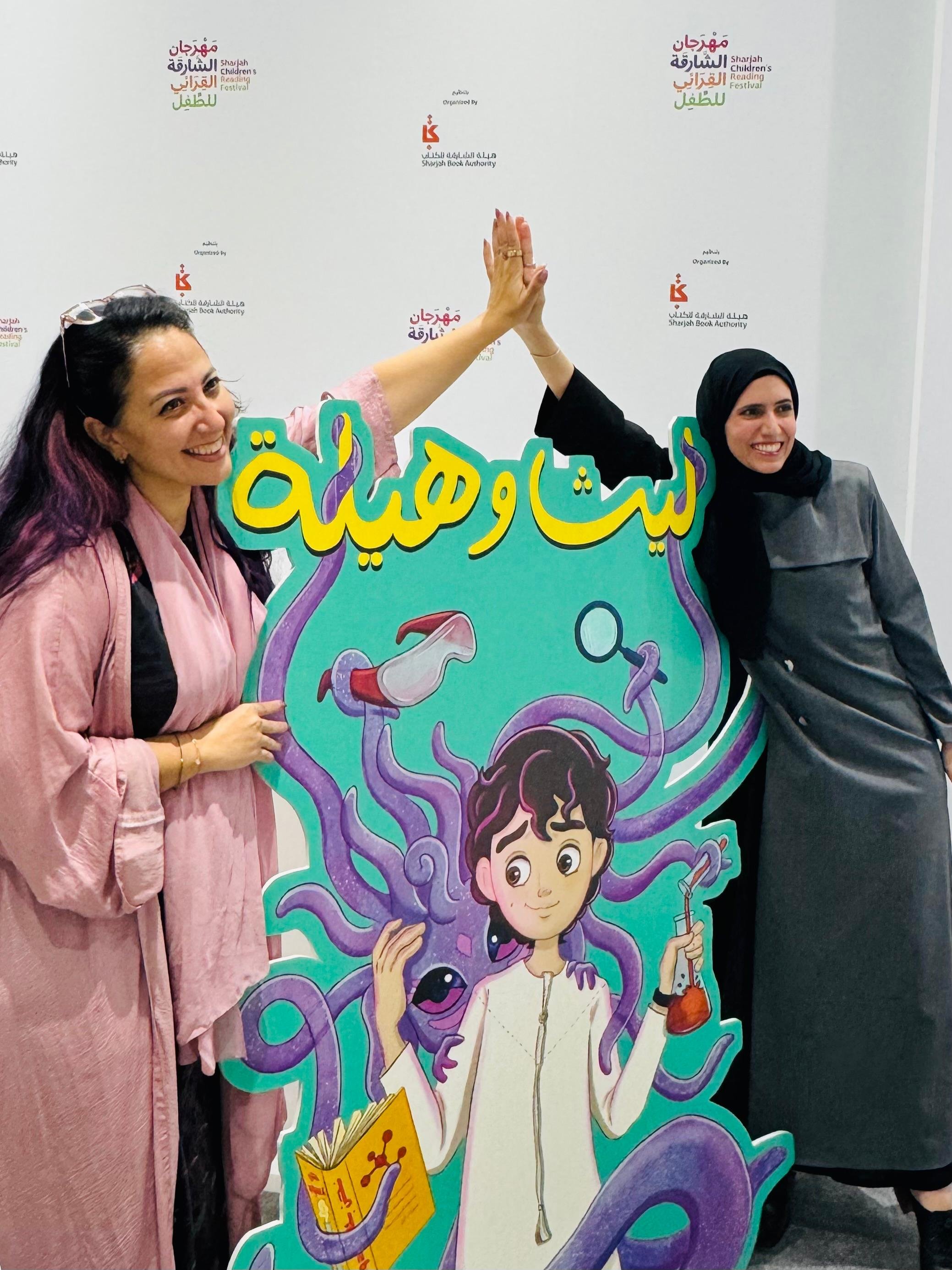 Two women are standing next to a colorful cartoon standee with an illustration of a girl with purple tentacles, holding a book and a test tube. The women are giving each other a high-five. The background has writing in Arabic and English indicating a