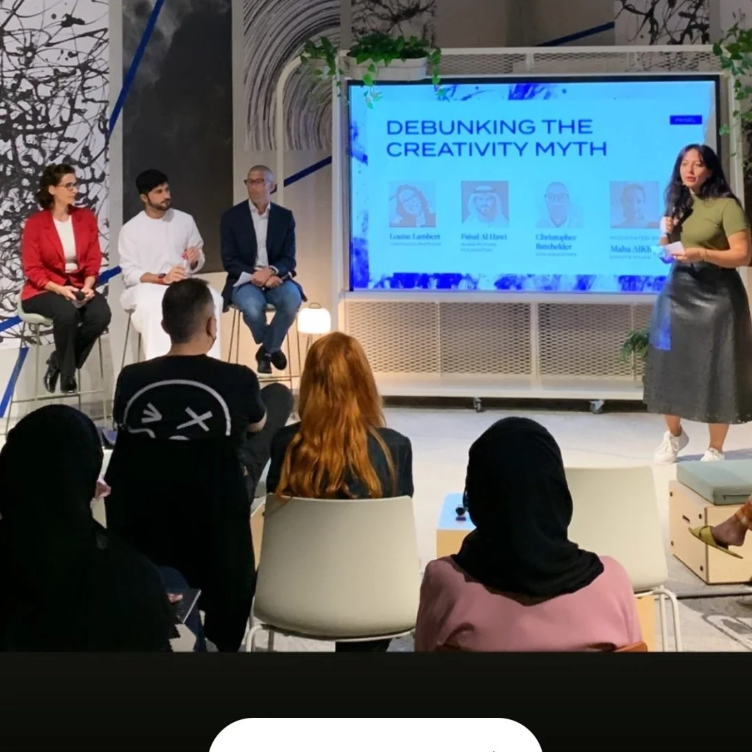 A panel discussion at a conference titled "Debunking the Creativity Myth." Four panelists sit on a stage, and a woman stands at the side, speaking into a microphone. The audience is seated facing the panel, with some taking notes. The background features a large screen displaying the title and images of the panelists.