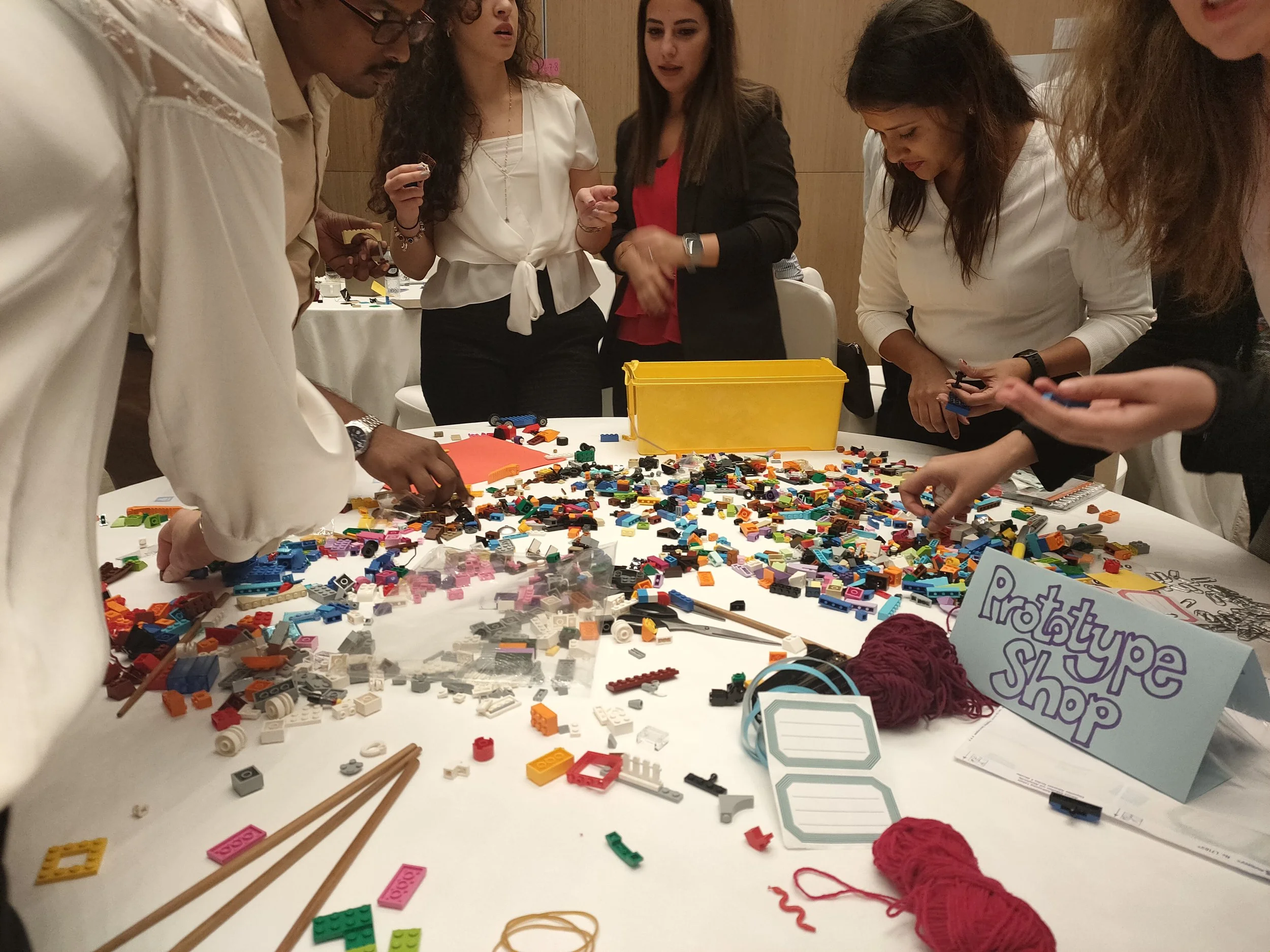 A group of people standing around a table covered with assorted LEGO pieces, some yarn, and notes, working on building their low-tech prototype to showcase their idea to the room.