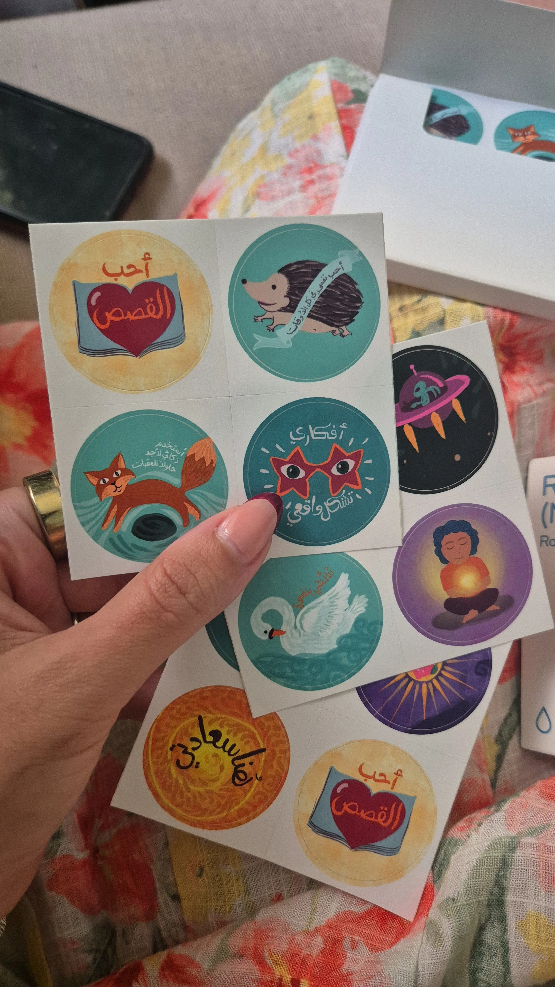 Sheet of colorful stickers with Arabic text and illustrations including a book with a heart, a hedgehog, a fox, star-shaped glasses, a spaceship, a swan, and a person meditating.