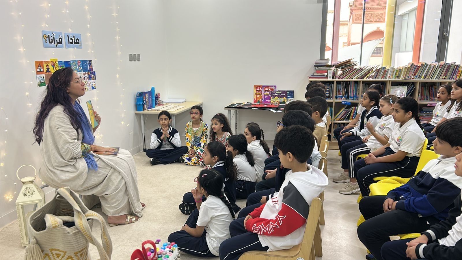 A woman reading a book to a group of children in a library, with bookshelves filled with books behind them and children sitting attentively on yellow and wooden chairs.