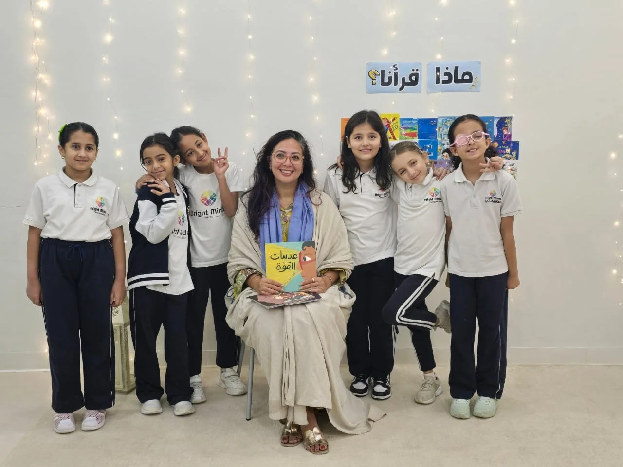 A class of seven young schoolgirls and their teacher posing together in a classroom, with some of the girls making peace signs. The teacher is seated in the center holding a book, and the girls are standing around her. The background features a white
