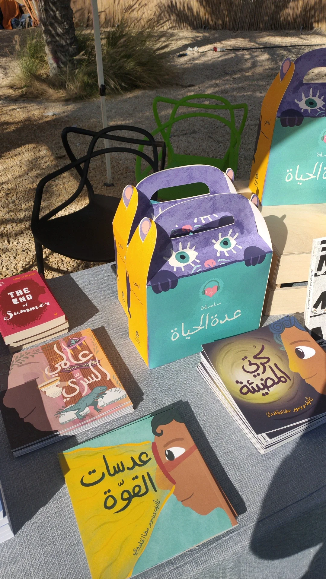 Table of books and gift bags with Arabic titles at an outdoor event, with plants and a wooden fence in the background.