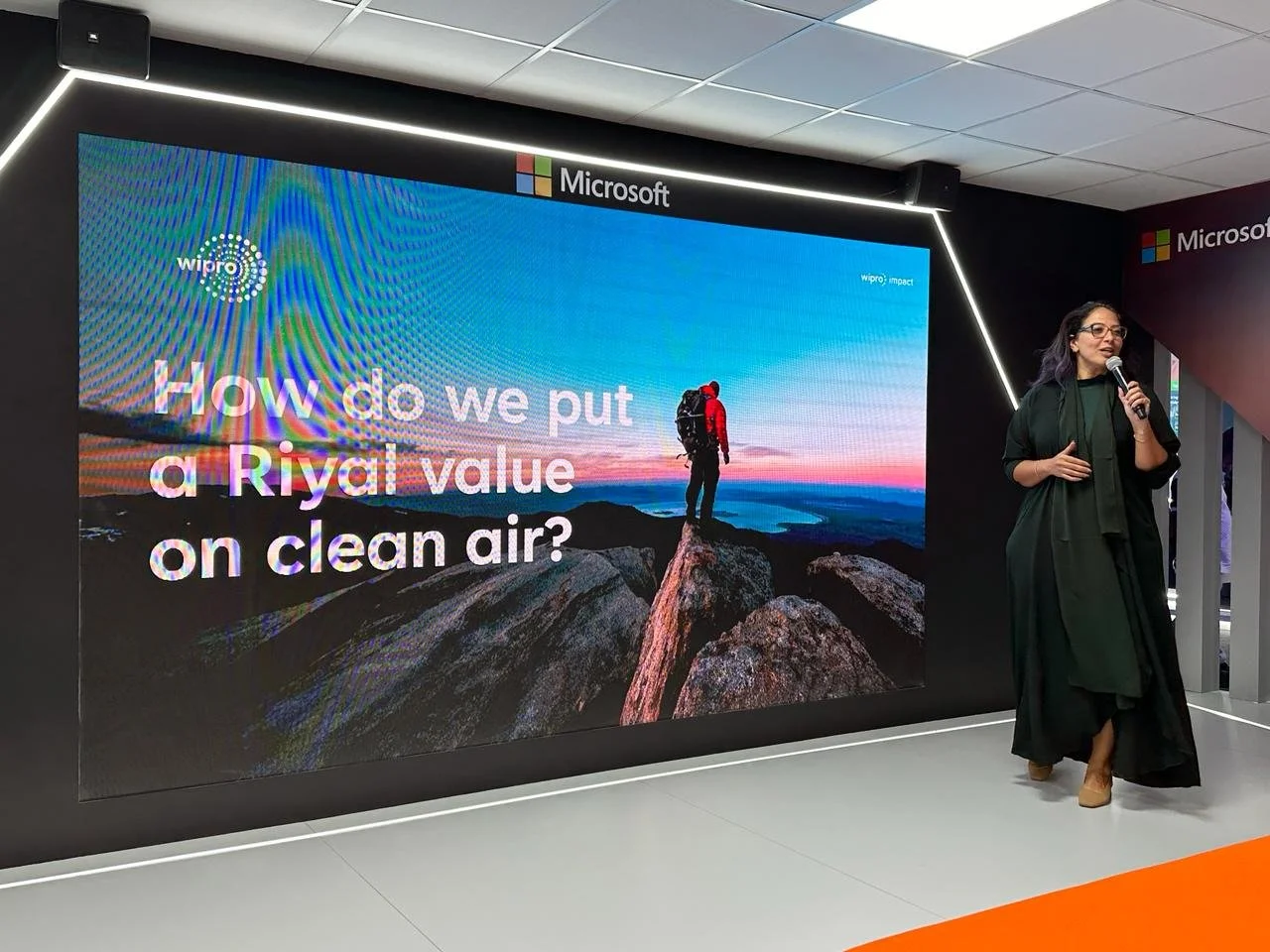 Maha in dark clothing speaking into a microphone next to a large screen at a Microsoft stage at LEAP 2024 in Riyadh, with a presentation slide showing a hiker on a mountain at sunset and the text: 'How do we put a Riyal value on clean air?'