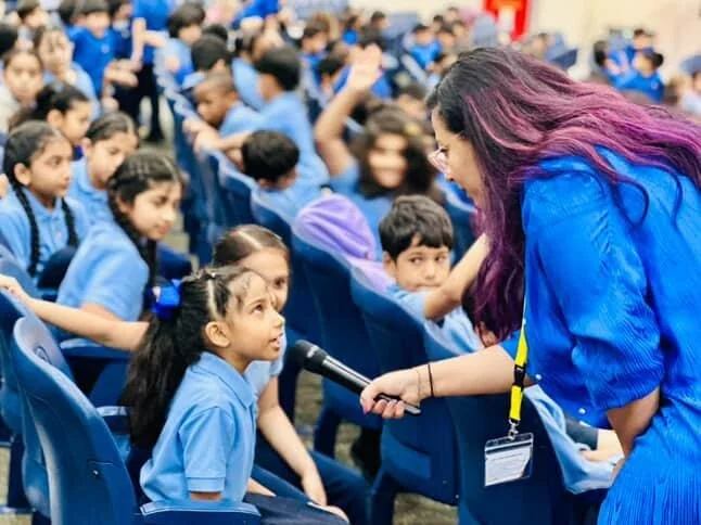 A woman with purple hair holding a microphone interacting with a young girl during an outdoor event with many children in blue uniforms sitting in rows.