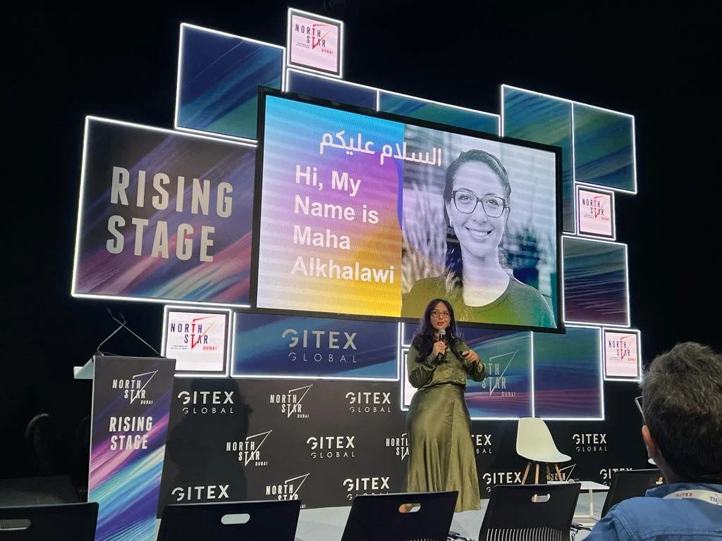 A woman on stage at GITEX Global in Dubai, speaking into a microphone. Behind her is a large screen displaying her name, Maha Alkhawali, in English and Arabic, with her photo. The stage banner reads 'Rising Stage' and 'North Star Dubai'.