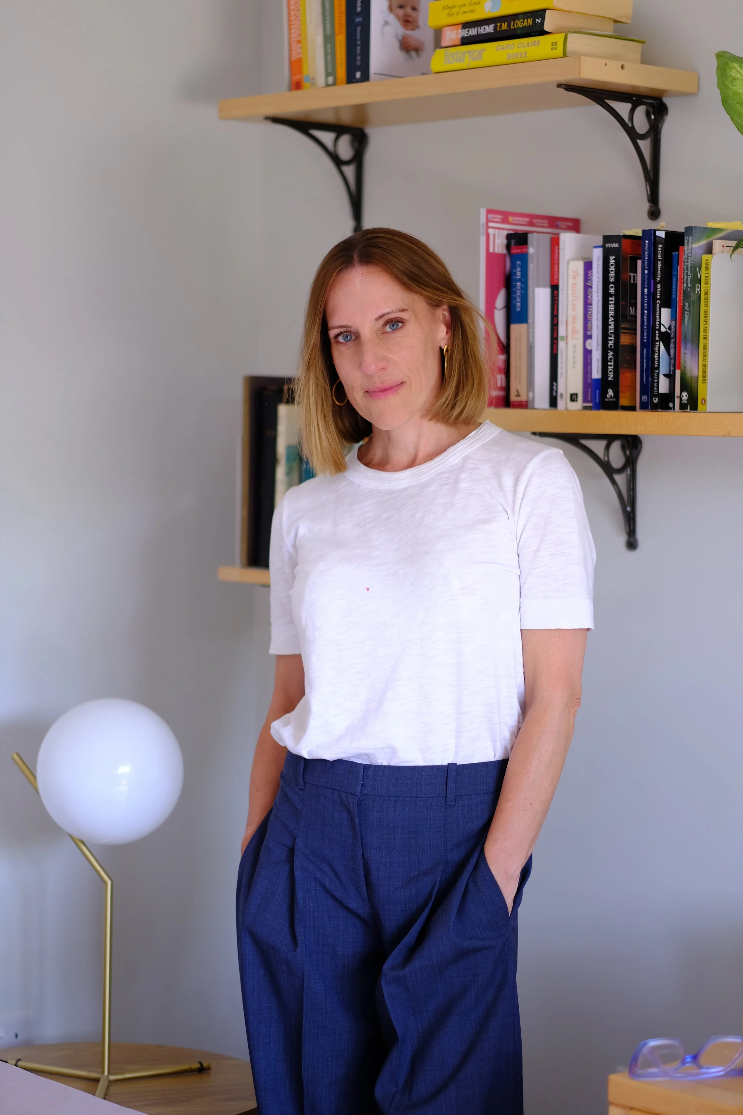 A woman with shoulder-length blonde hair wearing a white t-shirt and dark blue trousers standing in a room with a minimalist design. Behind her, there are floating wooden shelves with books, a modern lamp with white globes on a gold stand, and a pair of glasses on a wooden surface.