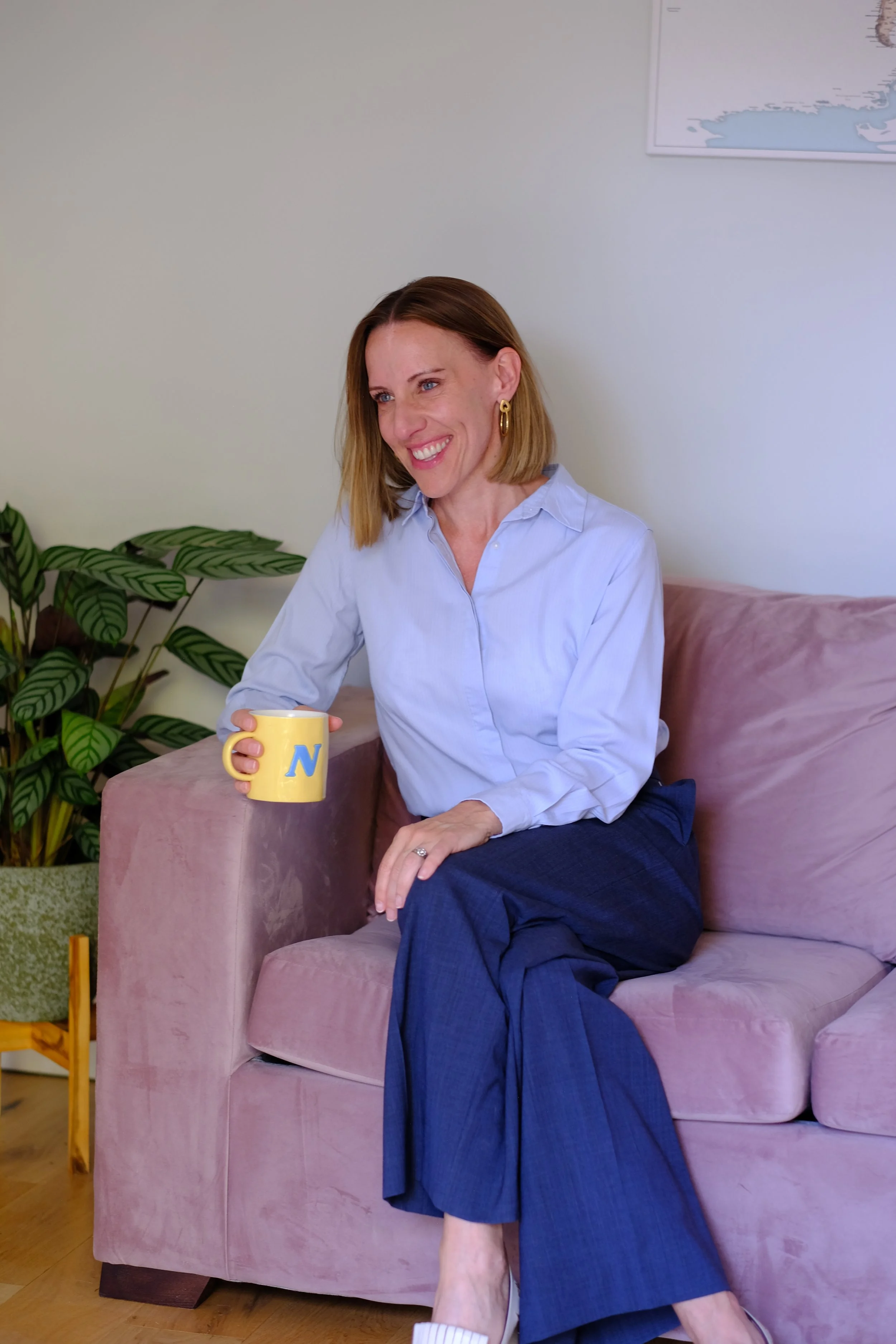 A woman with shoulder-length light brown hair sitting on a pink velvet couch, smiling while holding a yellow mug with blue text. She is wearing a light blue button-up shirt, dark blue pants, gold earrings, and white slippers, with a large green plant behind her and a map on the wall.
