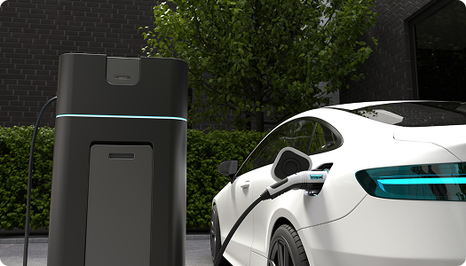 Electric car plugged into a charging station outdoors near a brick building and greenery.