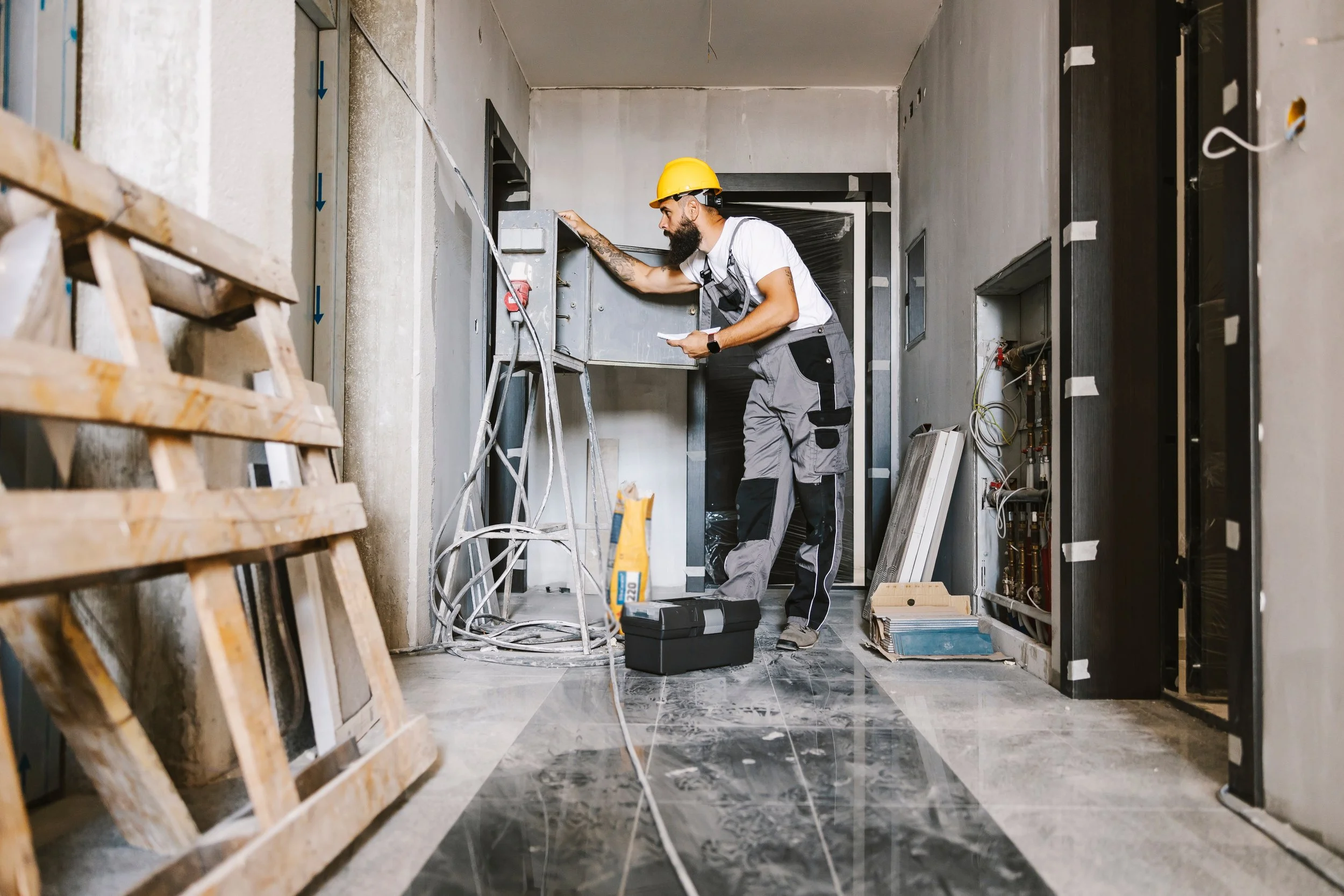 A man wearing a yellow construction helmet and work clothes is working inside a building under construction. He is checking or adjusting electrical wiring or components in an electrical panel. The space is unfinished with exposed wires, tools on the floor, and construction materials around.