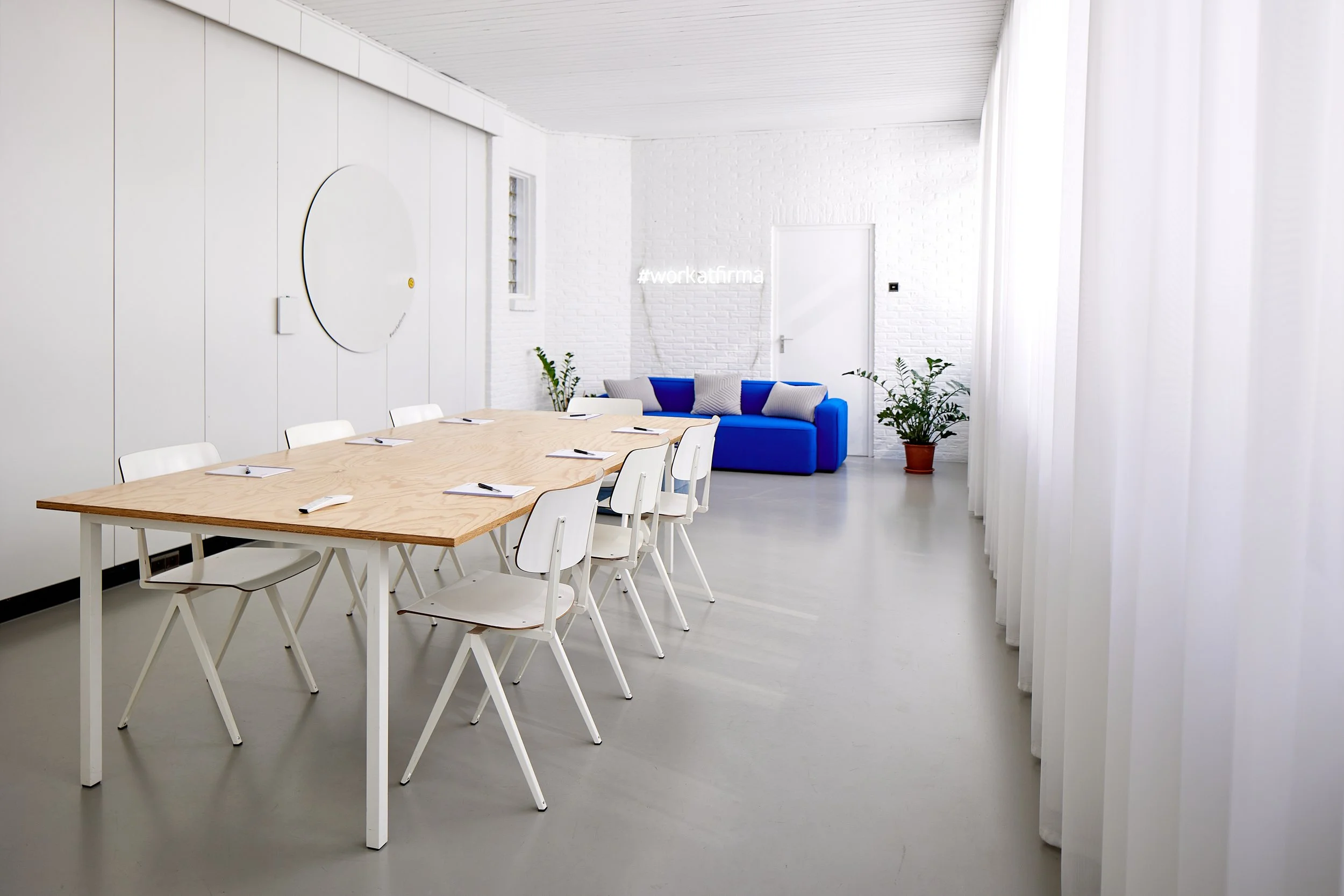 Een moderne vergaderruimte met een lange houten tafel, witte stoelen, een blauwe bank, planten en een hashtag '#workatfirma' op de muur.