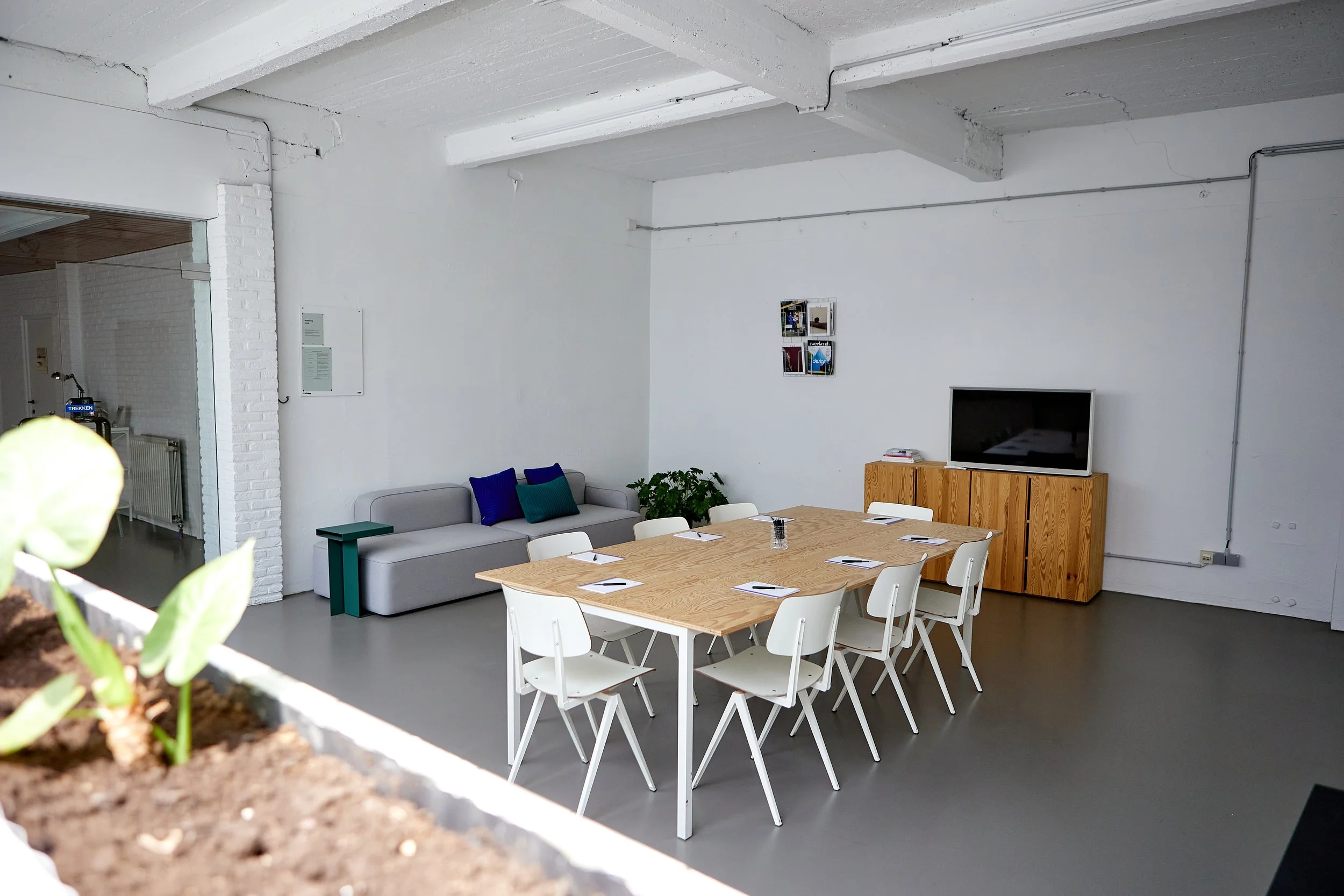 Binnenruimte met een grote houten tafel met witte stoelen, een beige bank met kussens, een vlakscherm tv op een houten kast, muurdecoraties en planten, in een sobere, industriële stijl met witte muren en plafond.