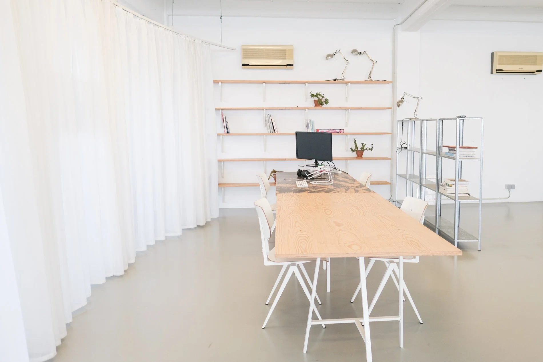Een minimalistisch kantoorruimtes met een houten tafel, witte stoelen, een metalen boekenrek met boeken, een computer en planten, witte muren, houten planken, en airconditioning.