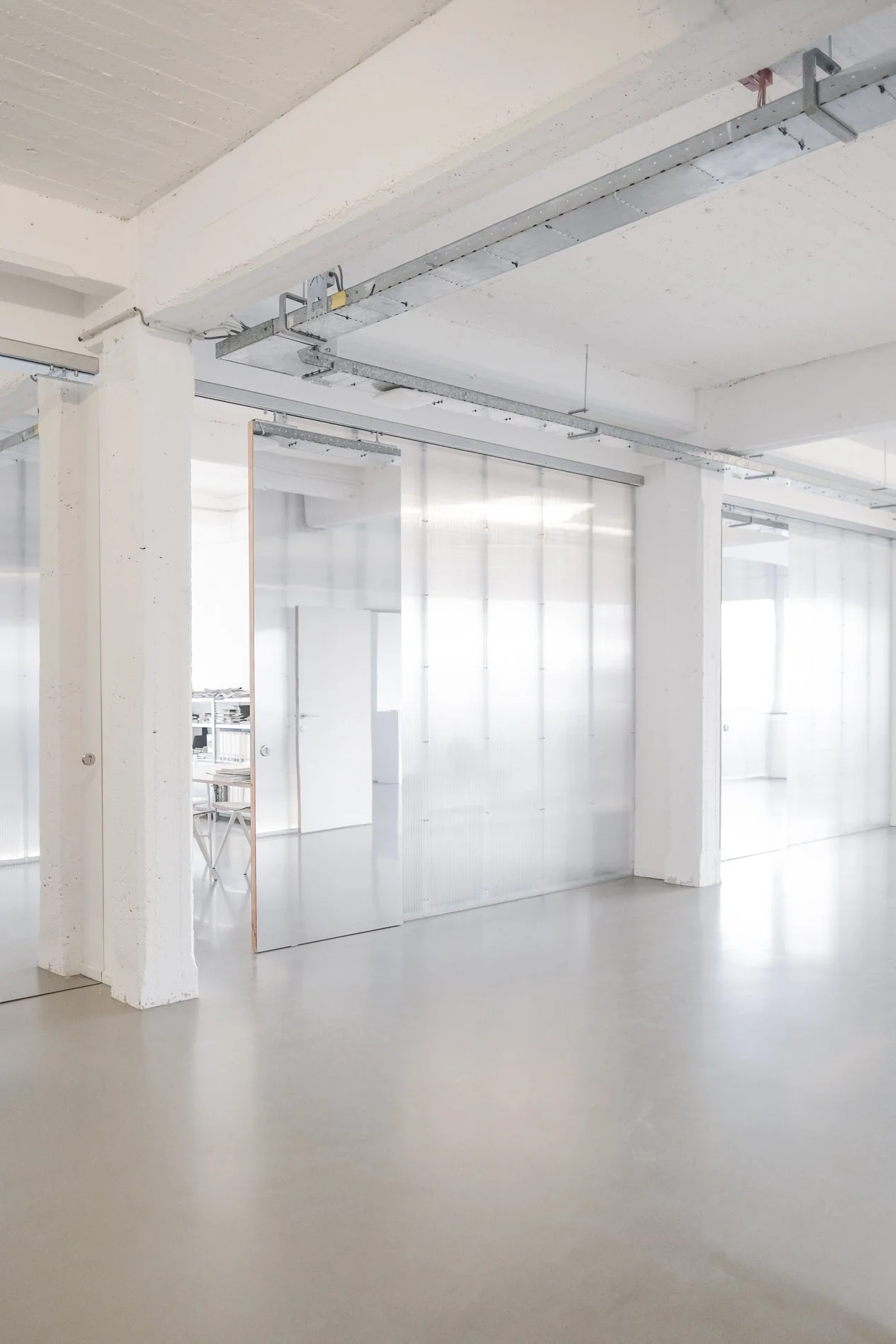 Een leeg kantoor met witte muren, plafond en vloer, doorzichtige scheidingswanden en een metalen installatierail aan het plafond.