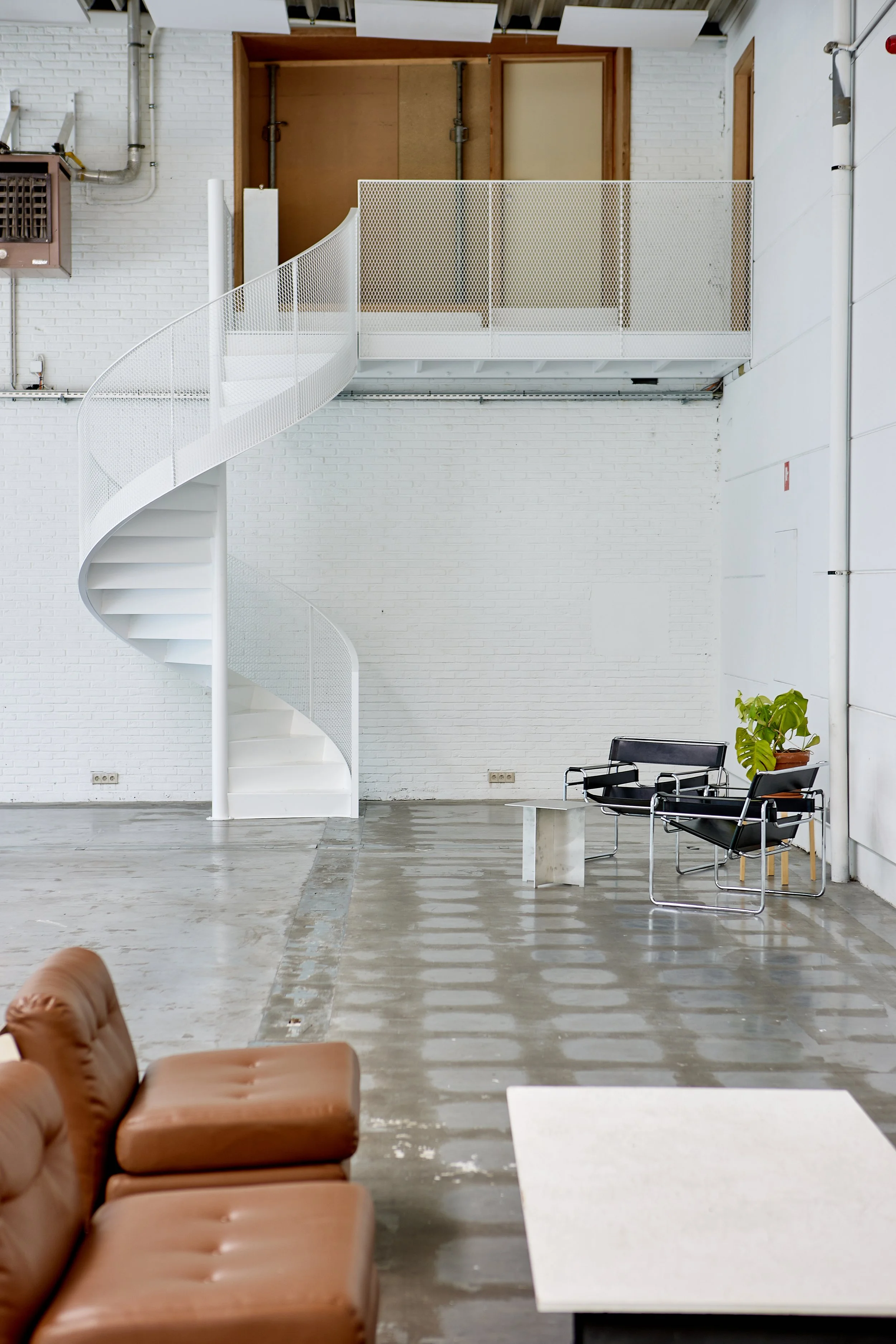 Interieur van een industrieel gebouw met een witte wenteltrap, een zitbank, een tafel, een plant, en een witgekalkte bakstenen muur.