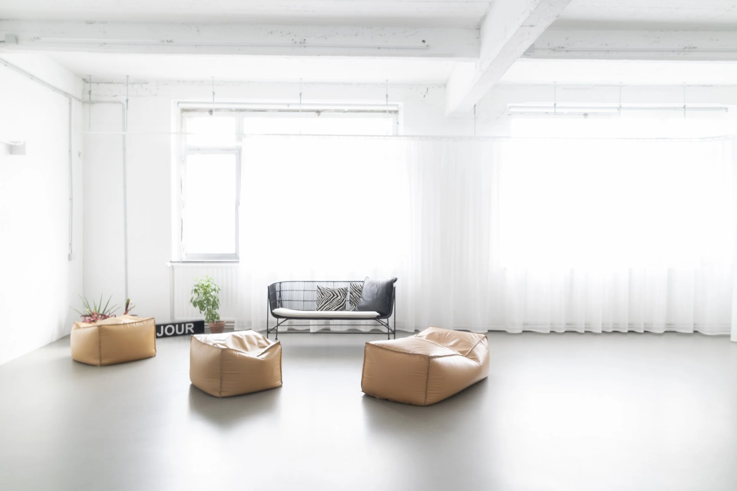 Een minimalistische woonkamer met veel natuurlijk licht, witte muren, een zwart metalen bank met decoratieve kussens, paar leren poefs in cognackleur, planten en een raam met witte gordijnen.