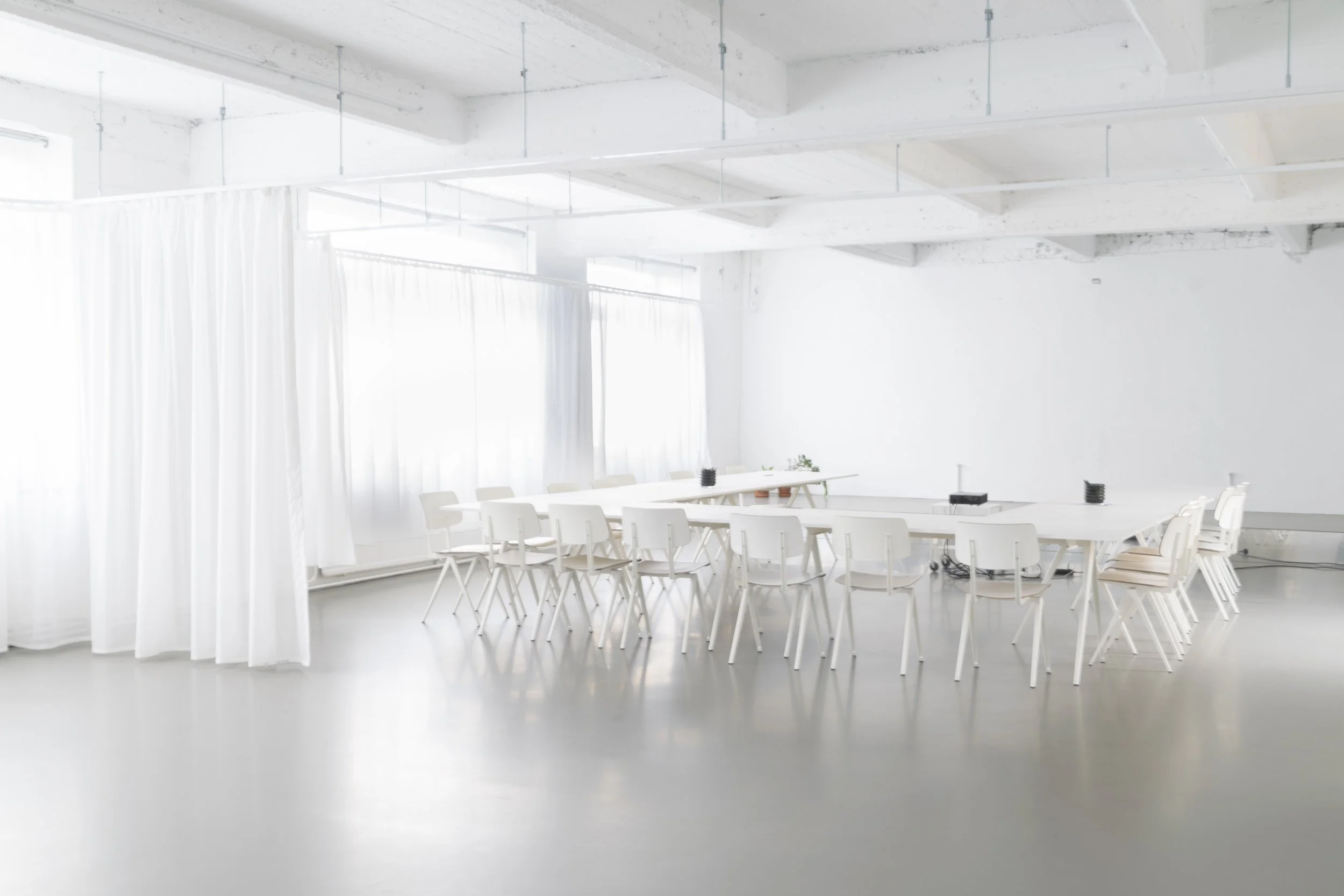 Een witte vergaderruimte met een lange tafel, witte stoelen en grote ramen met witte gordijnen.