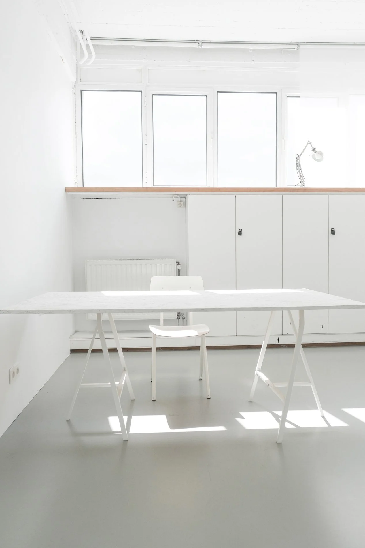 Minimalistisch wit interieur met een witte tafel en twee witte stoelen, grote ramen die veel licht binnenlaten, en een bureaulamp op de kast.