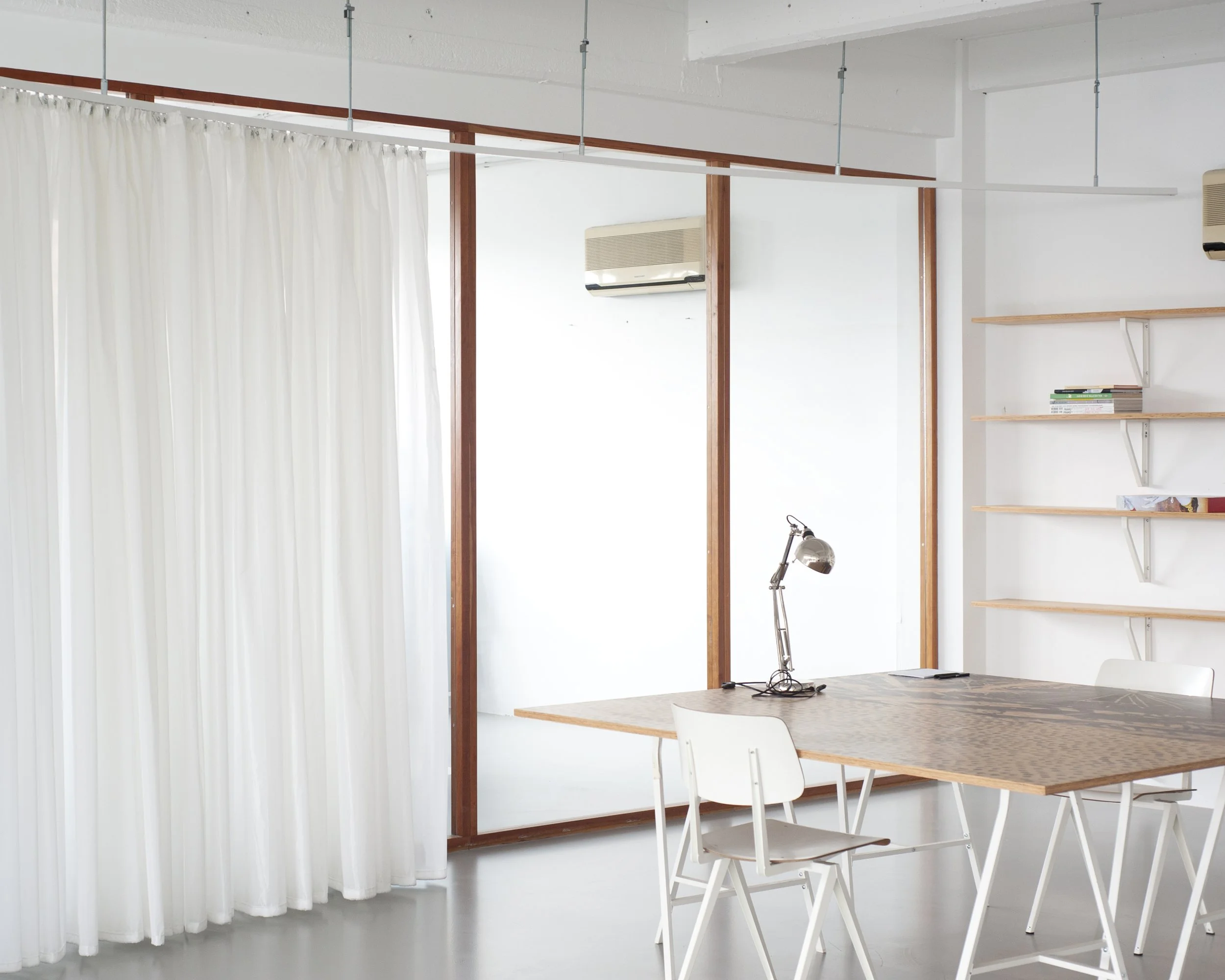 Een binnenruimte met witte muren, een houten tafel, witte stoelen, een boekenkast en een airconditioner aan de muur. Er hangen witte gordijnen.