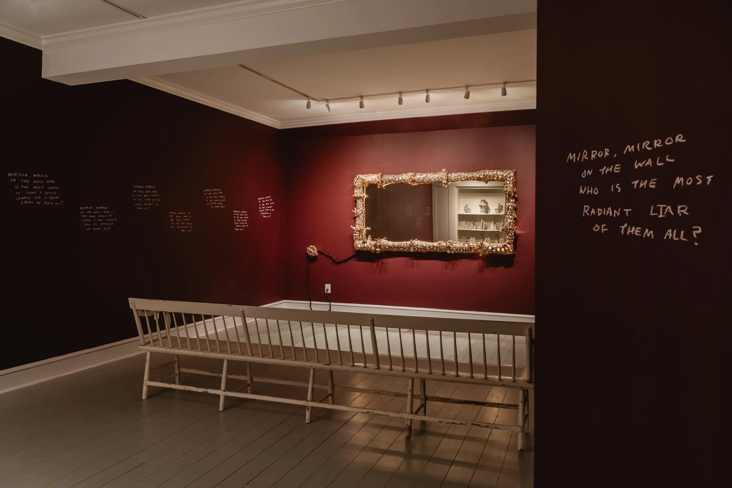 'Would an Angel Apologize for Spilling Champagne?' exhibition shot. To the right of the mirror, handwritten text on the wall reads, 'MIRROR, MIRROR ON THE WALL WHO IS THE MOST RADIANT LIAR OF THEM ALL?'