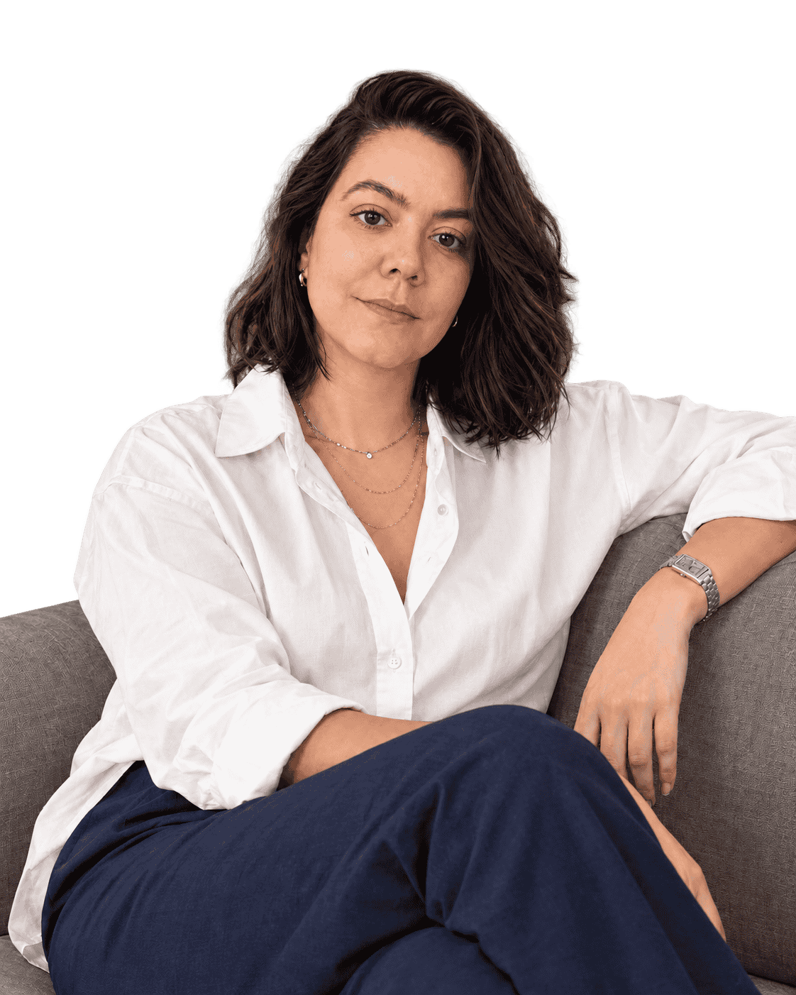 Mulher de cabelos castanhos sentada em um sofá cinza, usando camisa branca, calça azul-marinho, colares e relógio.