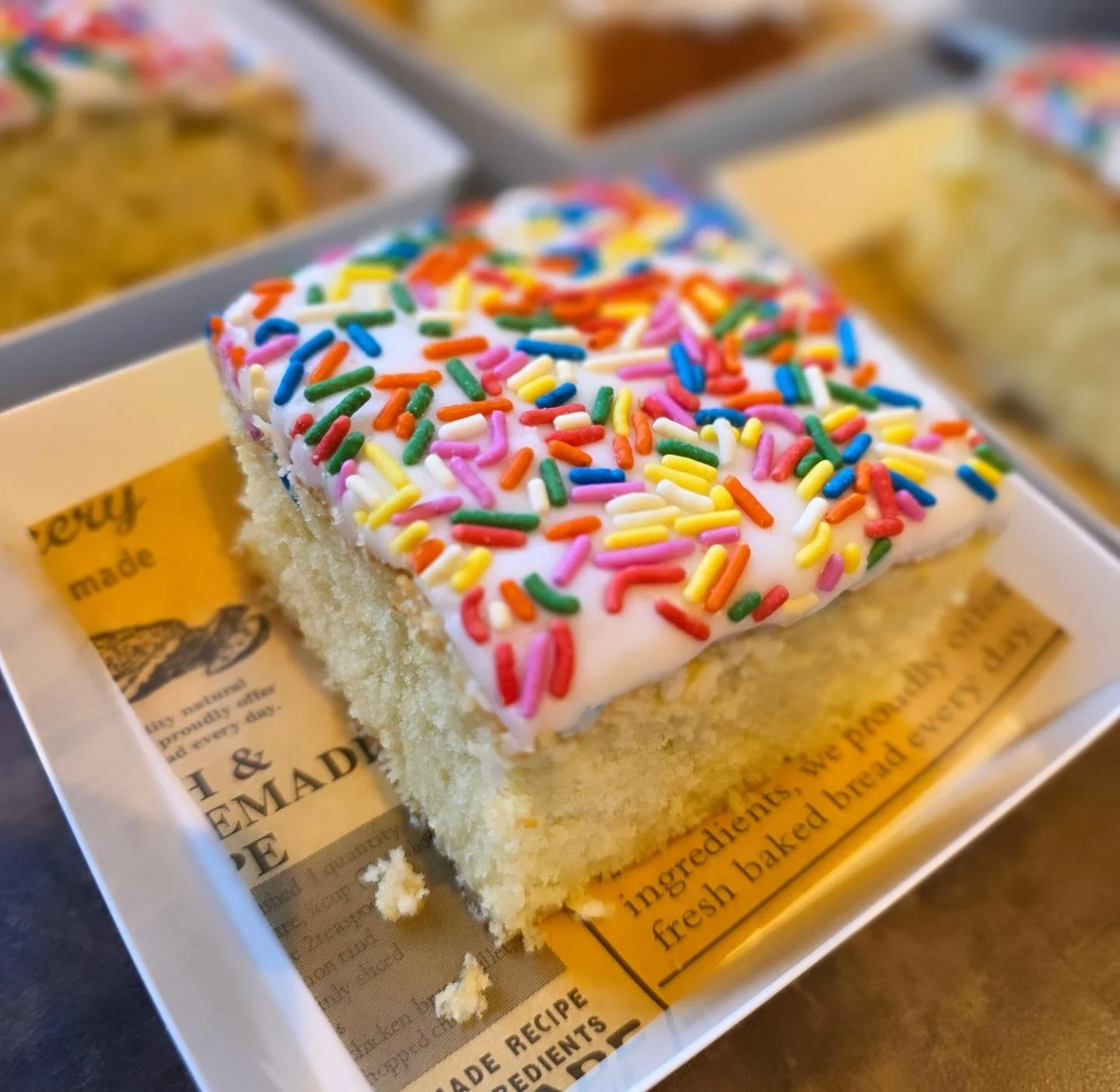 A square slice of vanilla cake with white frosting and colorful rainbow sprinkles, placed in a white box lined with paper that has printed text.