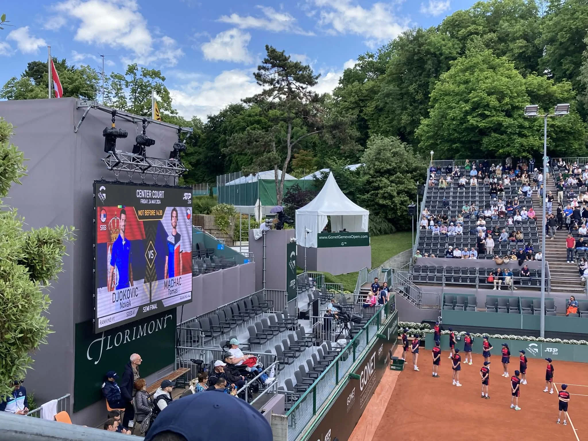 Spectators watching an ATP 250 tennis match at the Geneva Open, with the court and scoreboard displaying Djokovic versus Machac.