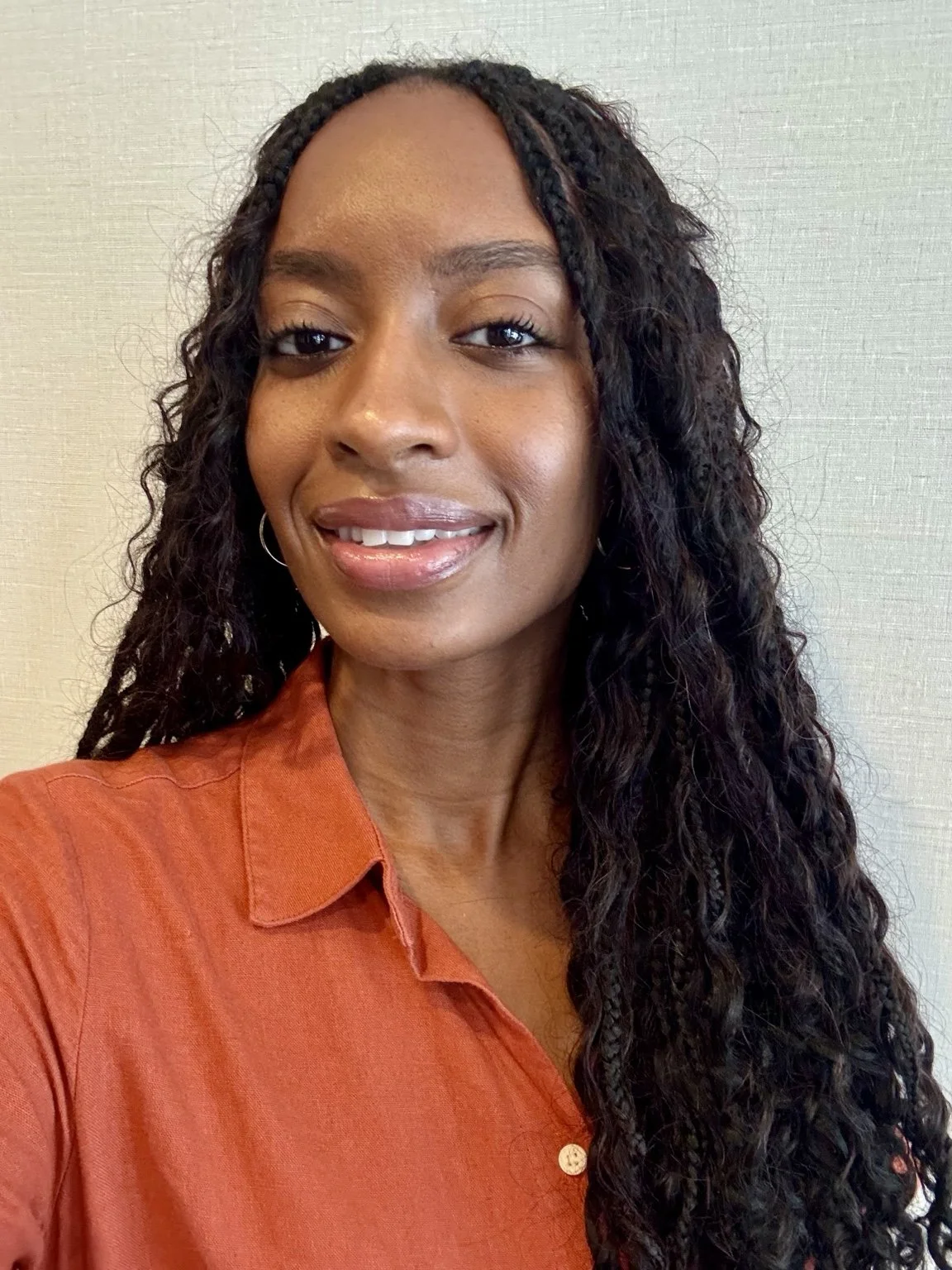 A woman with long curly black hair wearing an orange collared shirt, smiling, standing against a neutral background.
