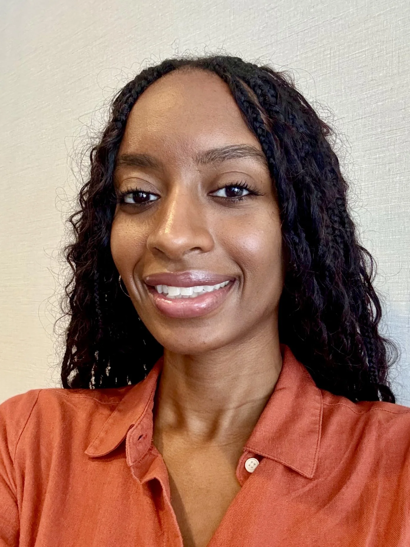A woman with medium skin tone and long curly black hair, smiling, wearing an orange shirt, standing against a light-colored wall.